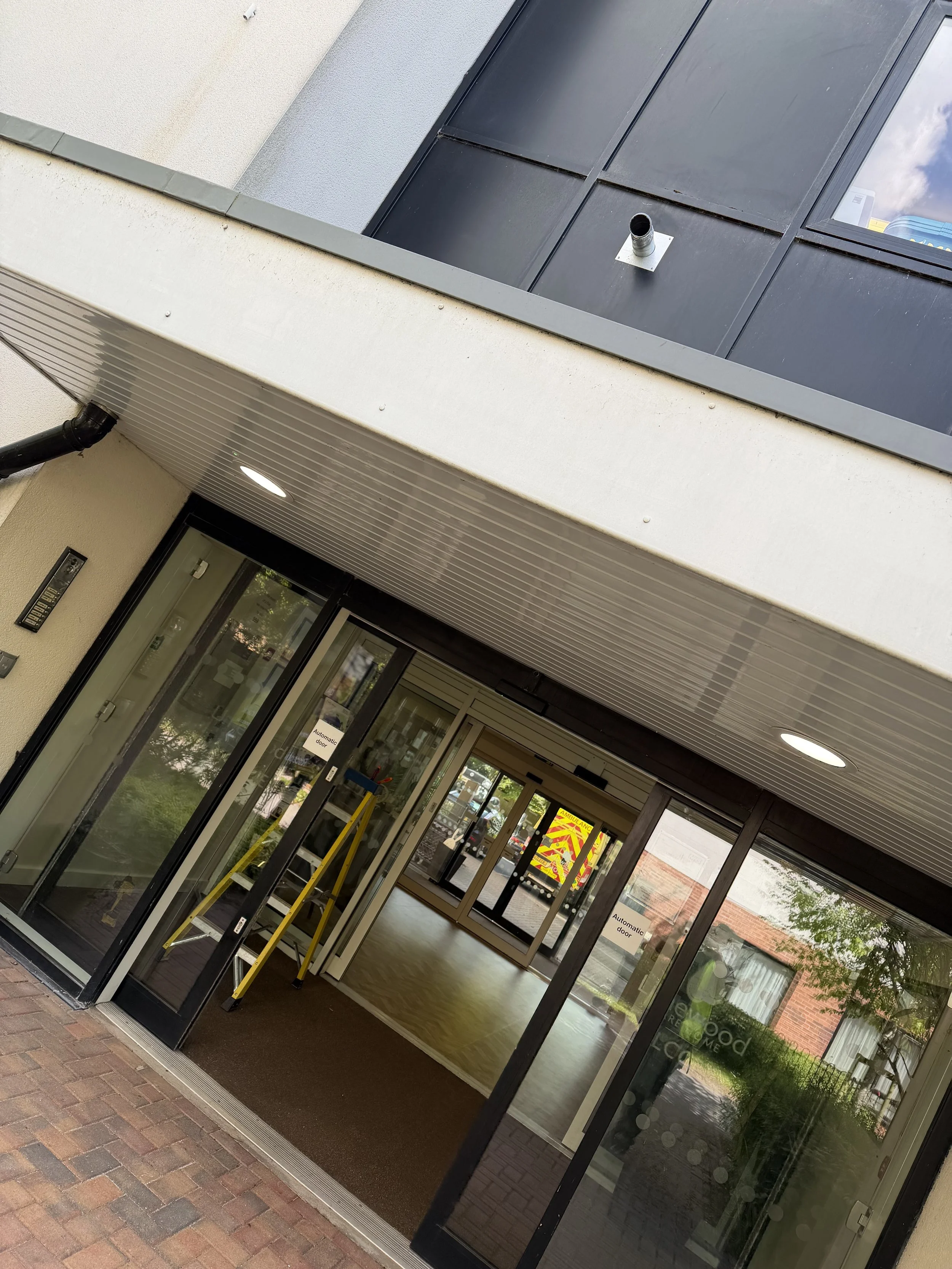 Entrance of a building with glass sliding doors, a brown mat, and a yellow caution sign inside visible through the glass. The ceiling above the entrance has recessed lighting. A sign on the door reads 'Automatic door'. Outside, there is a brick walkw