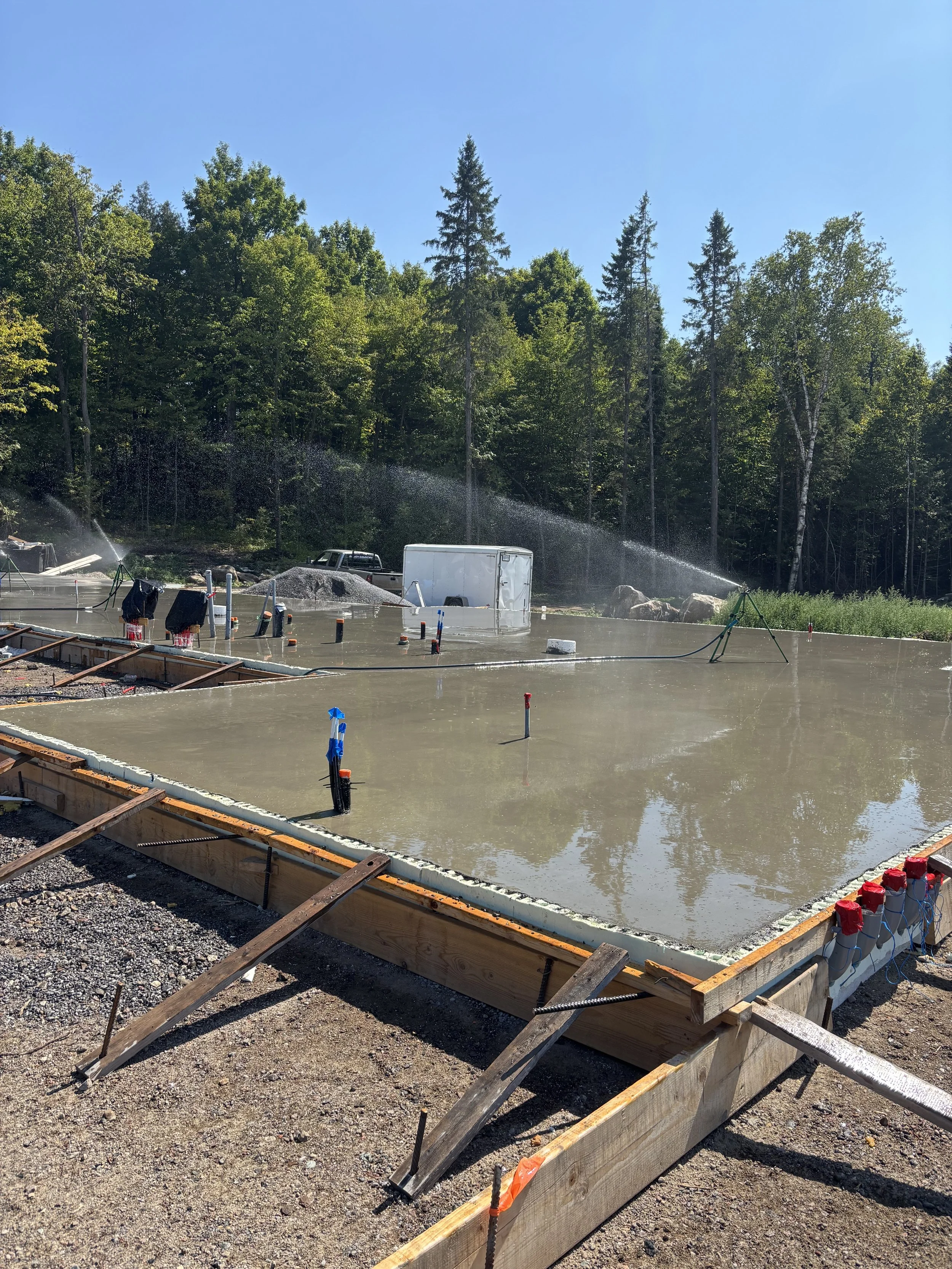 Construction site with freshly poured concrete slab, surrounded by wooden formwork, with tools and pipes, set against a backdrop of trees and blue sky.