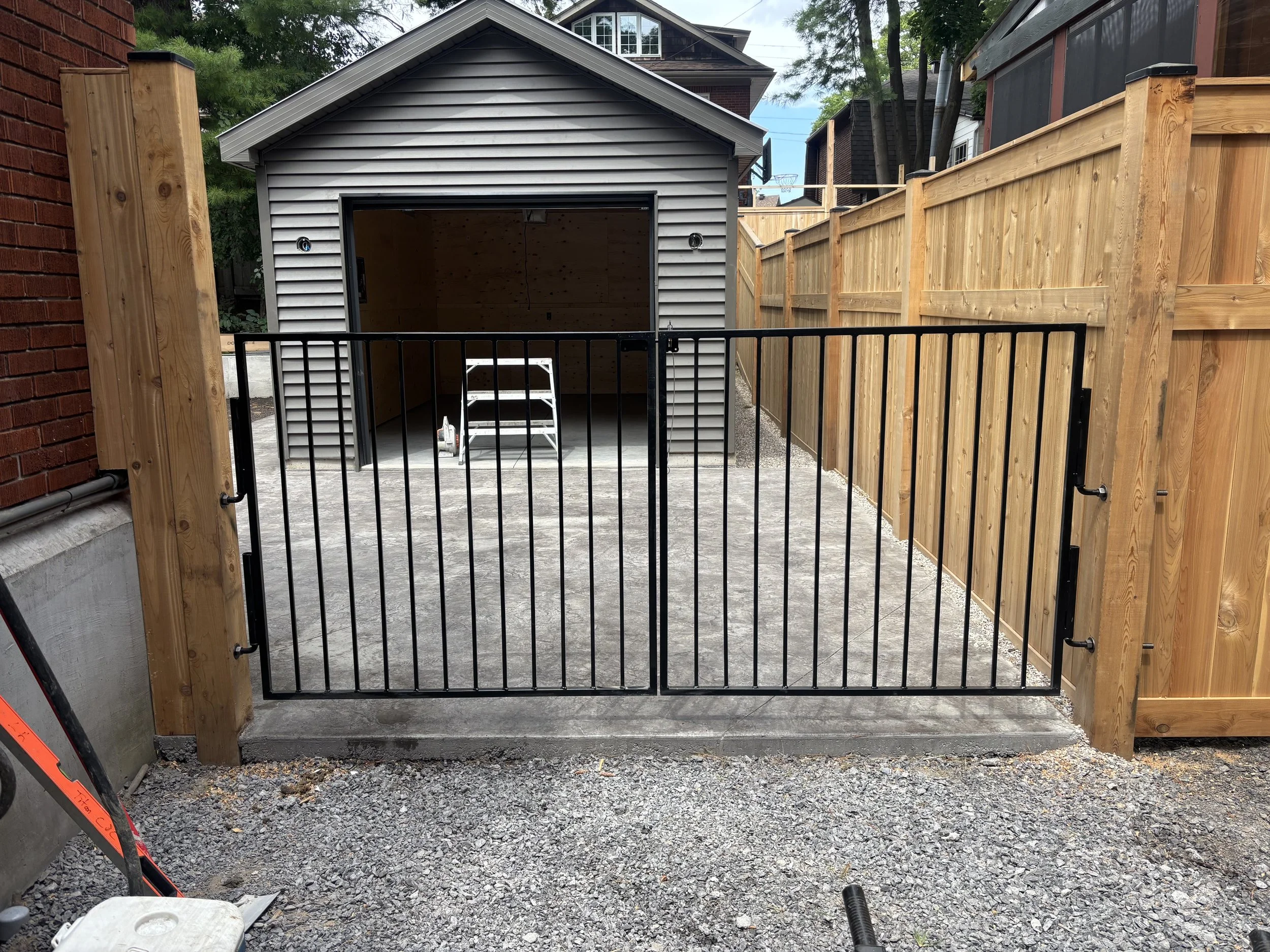 A black metal gate in front of a gravel driveway leading to a small gray shed with an open door, with a step ladder inside, enclosed by a wooden fence on either side.