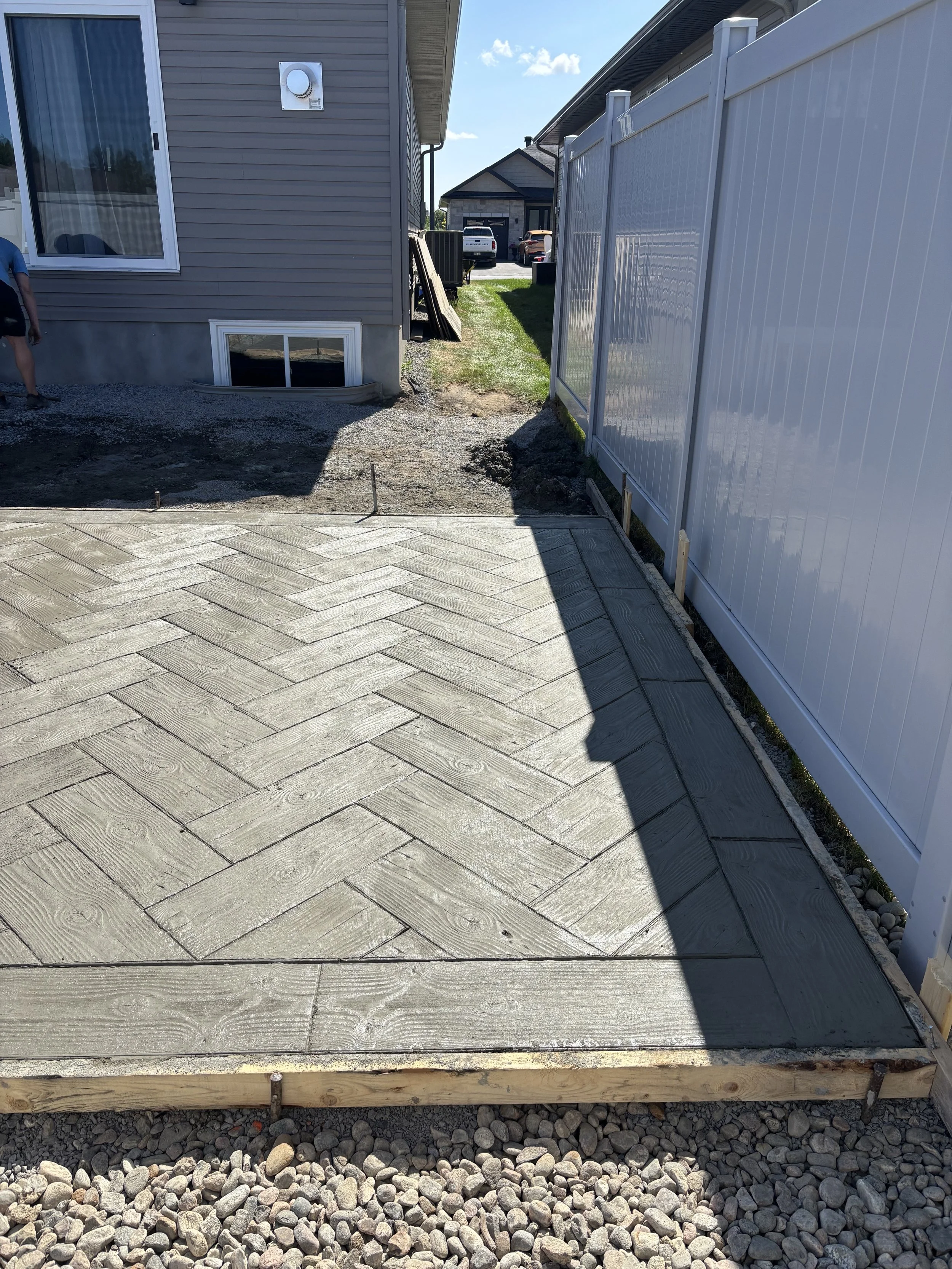 Recently laid concrete driveway with decorative herringbone pattern, new wooden border, gravel base, and white privacy fence on the right side.