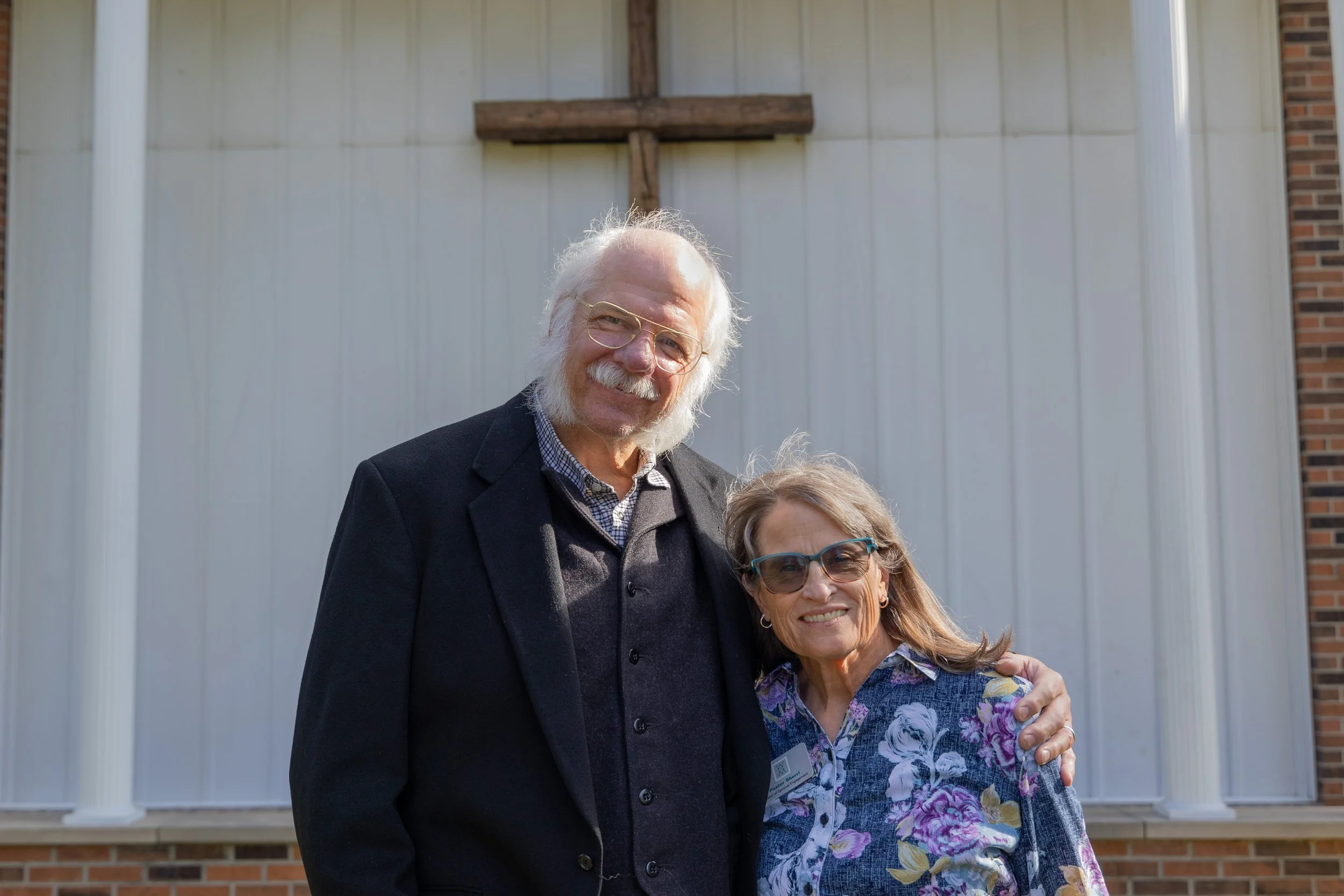 Older man with white hair and beard wearing suit jacket and glasses, and woman with long hair and glasses wearing floral blouse, smiling and standing close together outside in front of a white wall with a cross above.