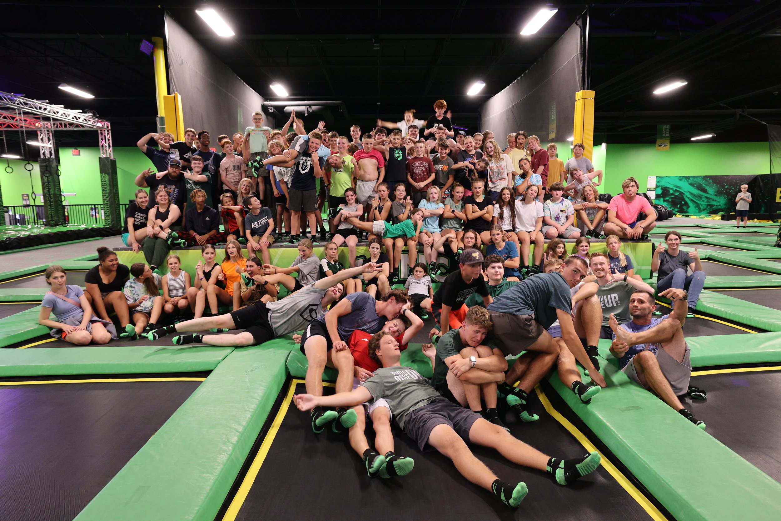 Large group of children and teenagers at an indoor trampoline park, some sitting, some standing, and some lying on the trampoline mats, with a few adults among them, in a colorful, energetic setting.