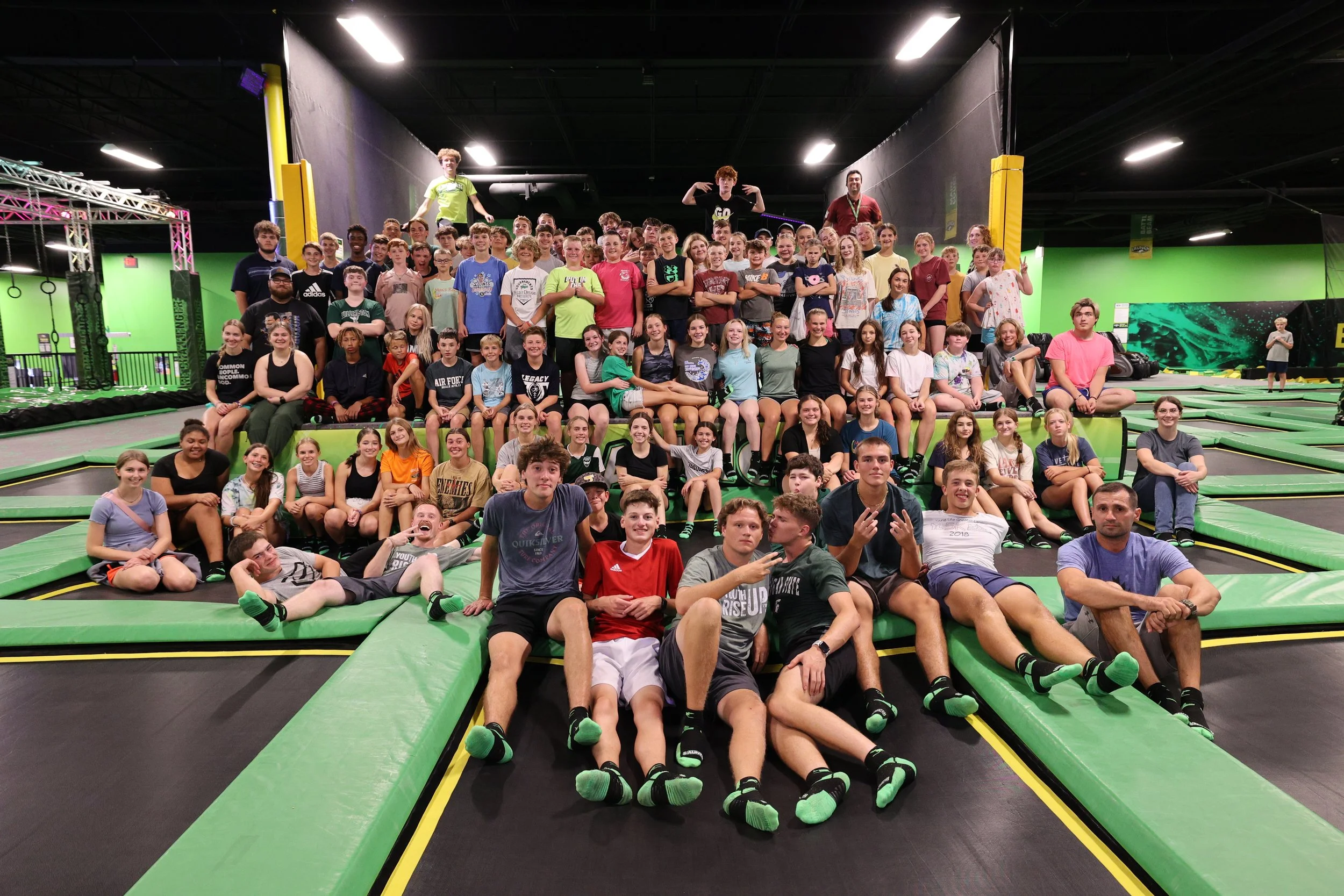 Large group of children and teenagers gathered at an indoor trampoline park, sitting and standing on the trampolines, with some on the padded steps and others on the trampoline areas, smiling and posing for the photo.