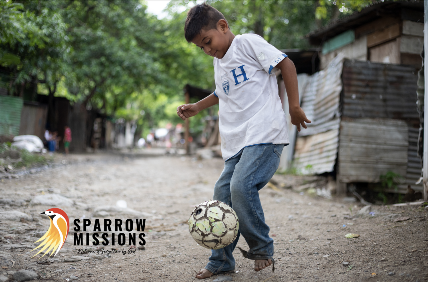 Young boy barefoot, playing soccer with a makeshift ball made of worn-out material, on a dirt pathway in a simple neighborhood with trees and modest houses.