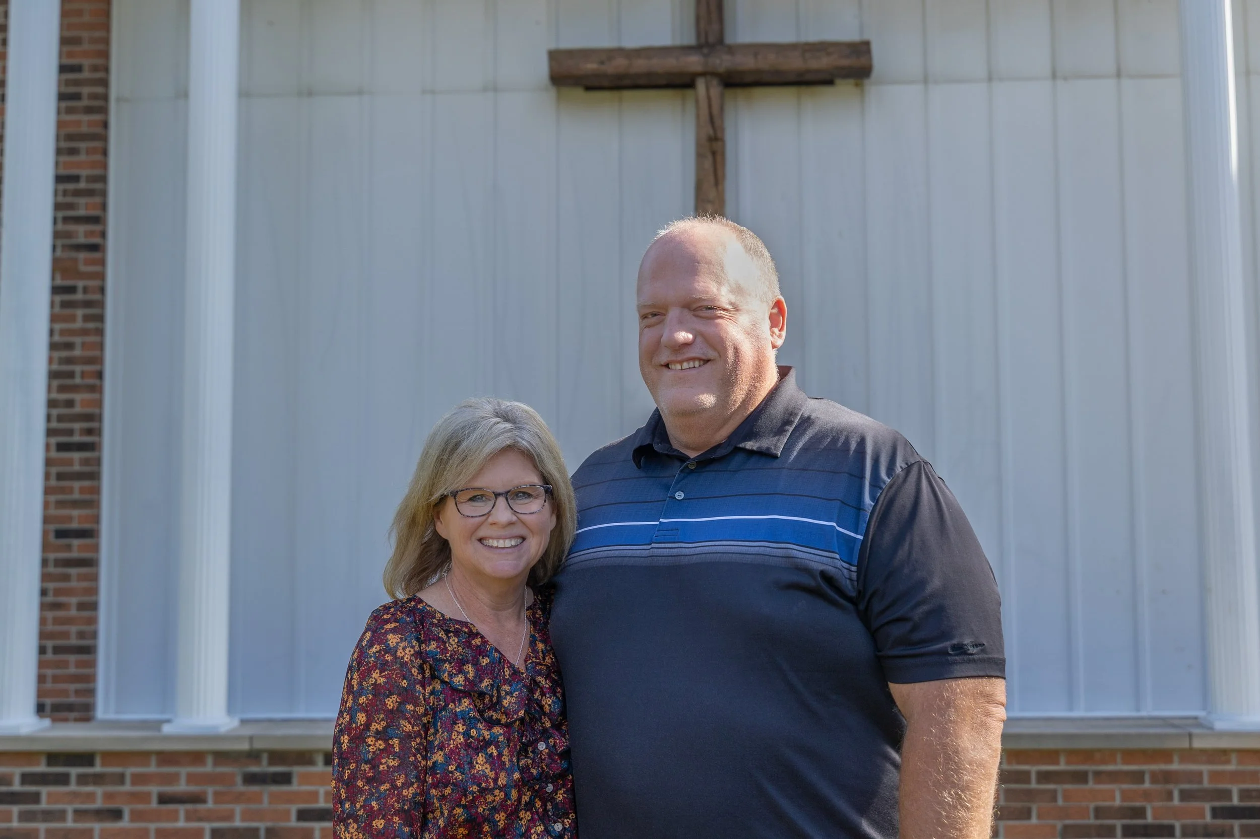 A smiling woman with glasses and a floral blouse standing next to a smiling man in a striped polo shirt, in front of a building with white siding, brick, and a wooden cross mounted on the wall.