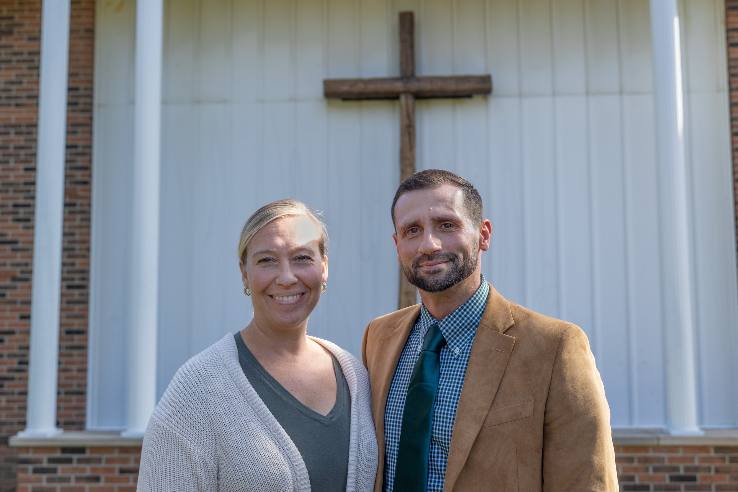 A man and woman standing in front of a church with a large wooden cross on the wall behind them. The woman has blonde hair and is smiling, wearing a light gray cardigan and dark gray top. The man has dark hair and a beard, wearing a tan blazer, blue checkered shirt, and blue tie.