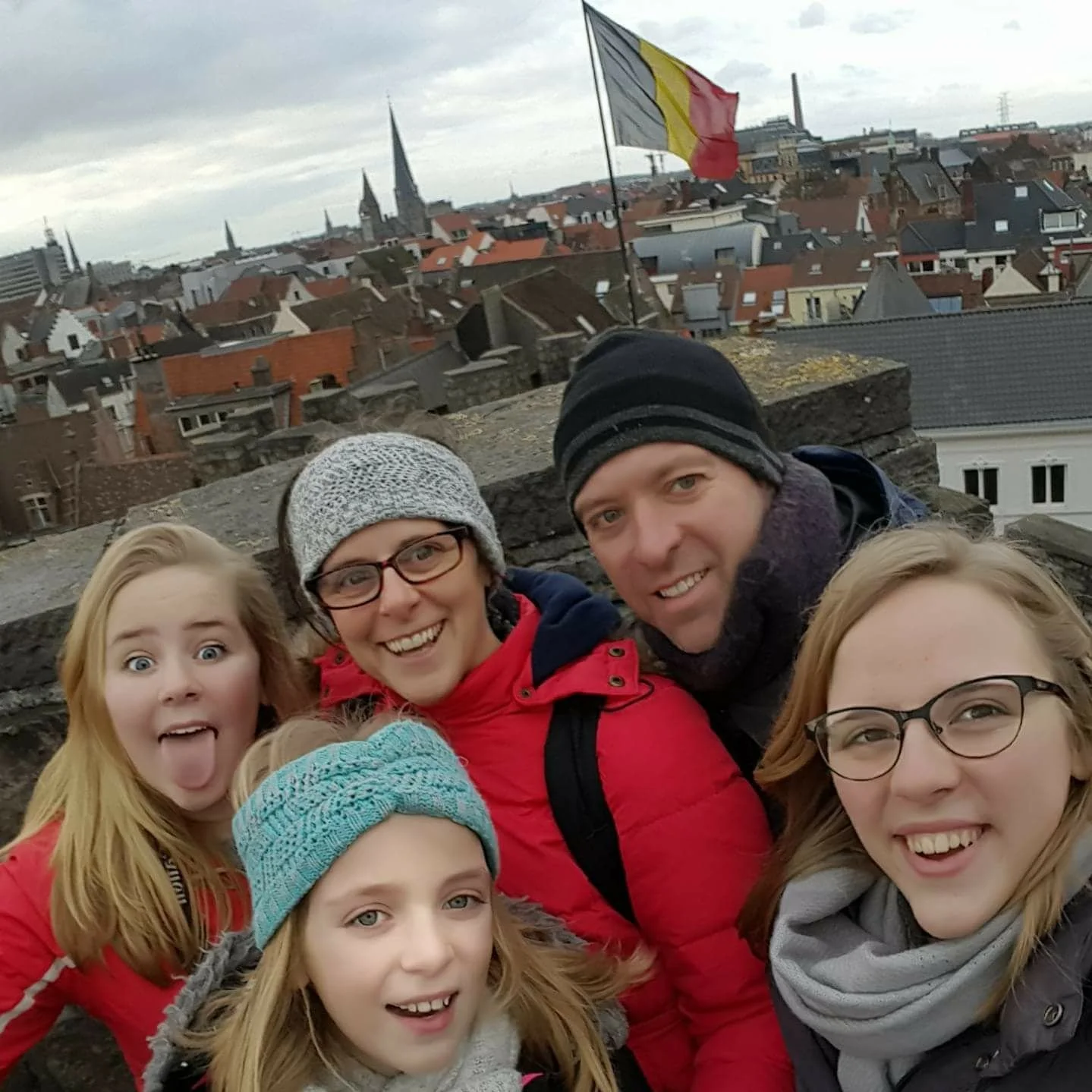 Group of six people, two children and four adults, smiling and posing for a selfie outdoors on a rooftop in a city with historic buildings and a Belgian flag in the background.
