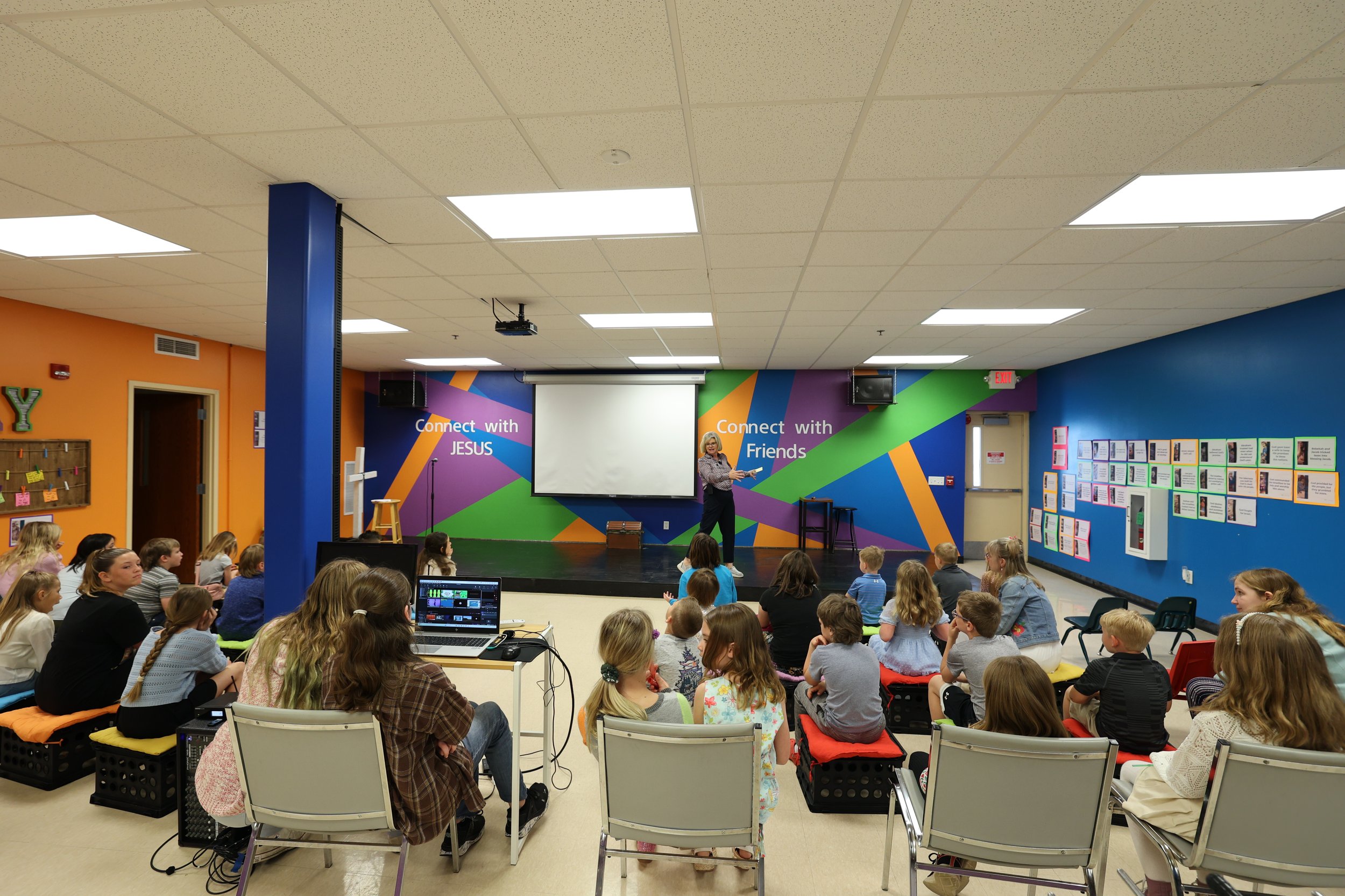 A woman gives a presentation to children seated on cushions in a colorful room with bright walls and motivational phrases.