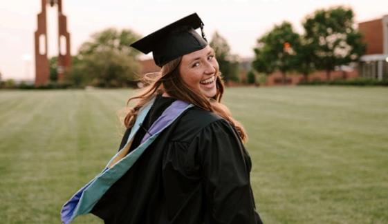 A young woman in a graduation cap and gown smiling outdoors on a grassy field with trees and a building in the background.