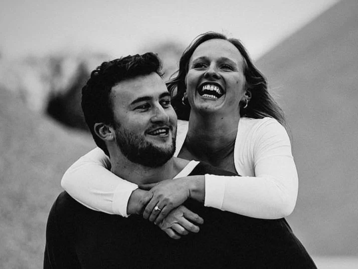 A happy couple, a man and a woman, laughing together outdoors, with the woman giving the man a piggyback ride.