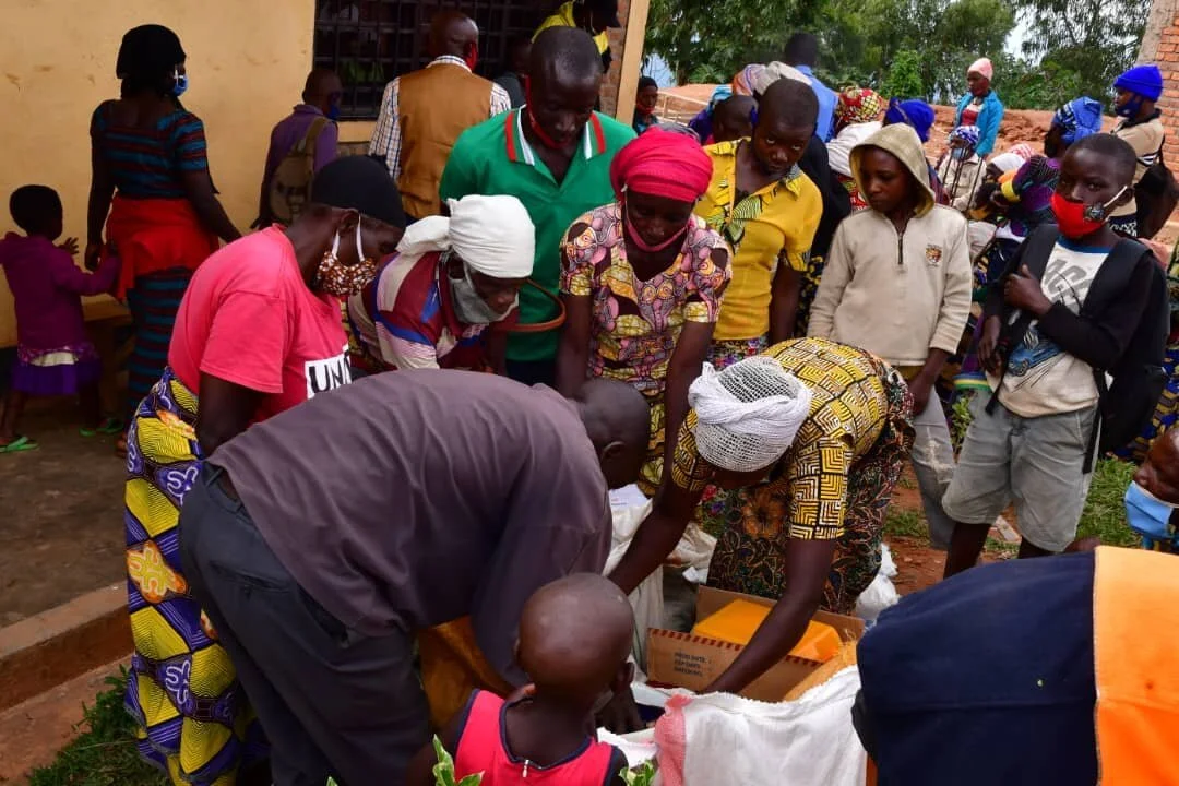 A group of people, including women and children, gathered outdoors around a table or box, engaging in a distribution or aid activity. Some individuals are wearing face masks, and the setting appears to be a community event or aid distribution.