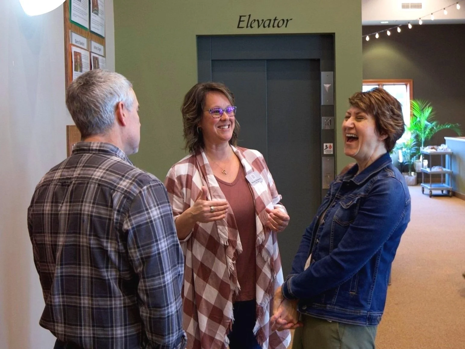 Four young women sitting at a table in a coffee shop, smiling and engaging with each other, using electronic devices and holding coffee cups.