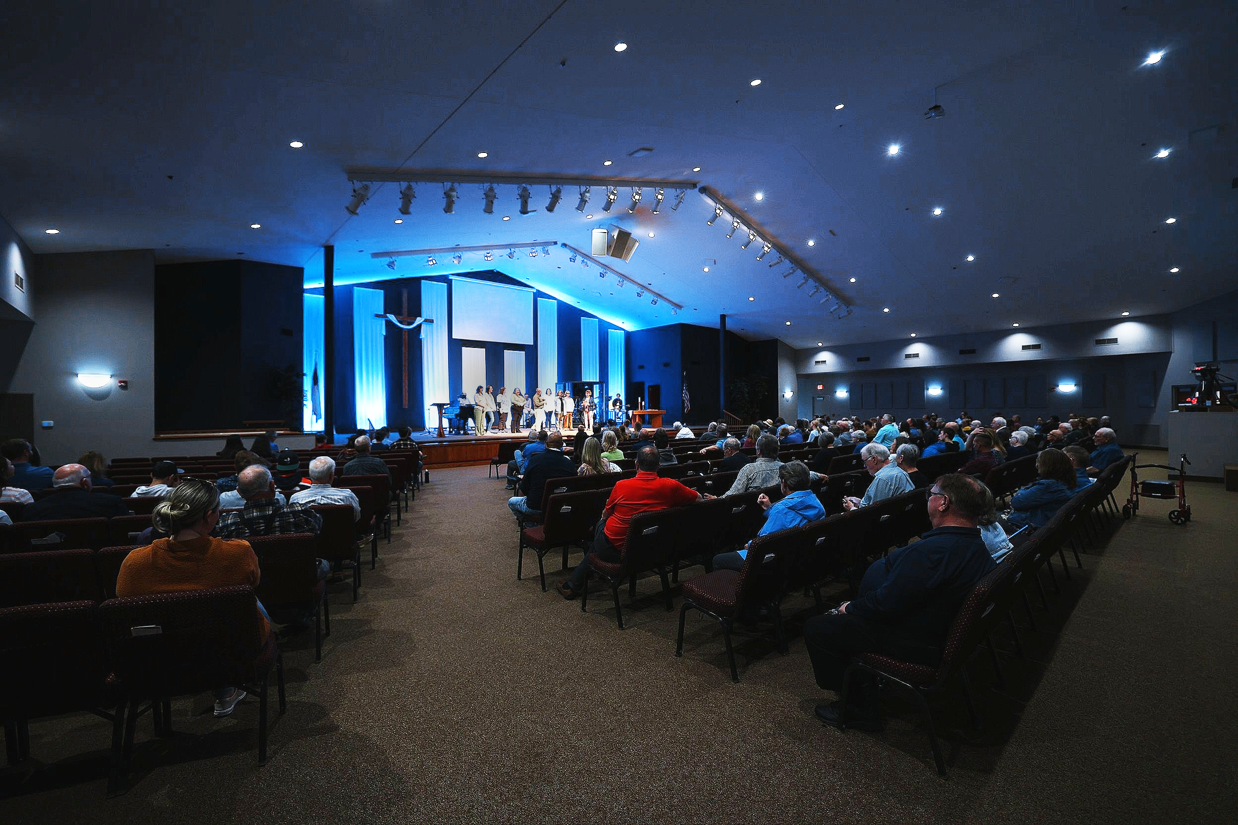 A large congregation attending a church service or event in a spacious auditorium with a stage illuminated in blue light, a cross hanging on the wall behind the stage, and a group of people singing or speaking.