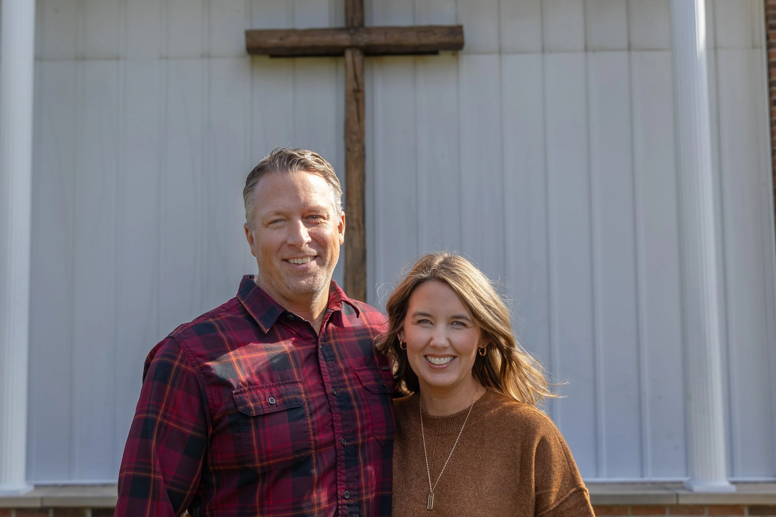 A smiling man and woman standing outdoors in front of a metal building with columns, the man wearing a red plaid shirt and the woman wearing a brown sweater.