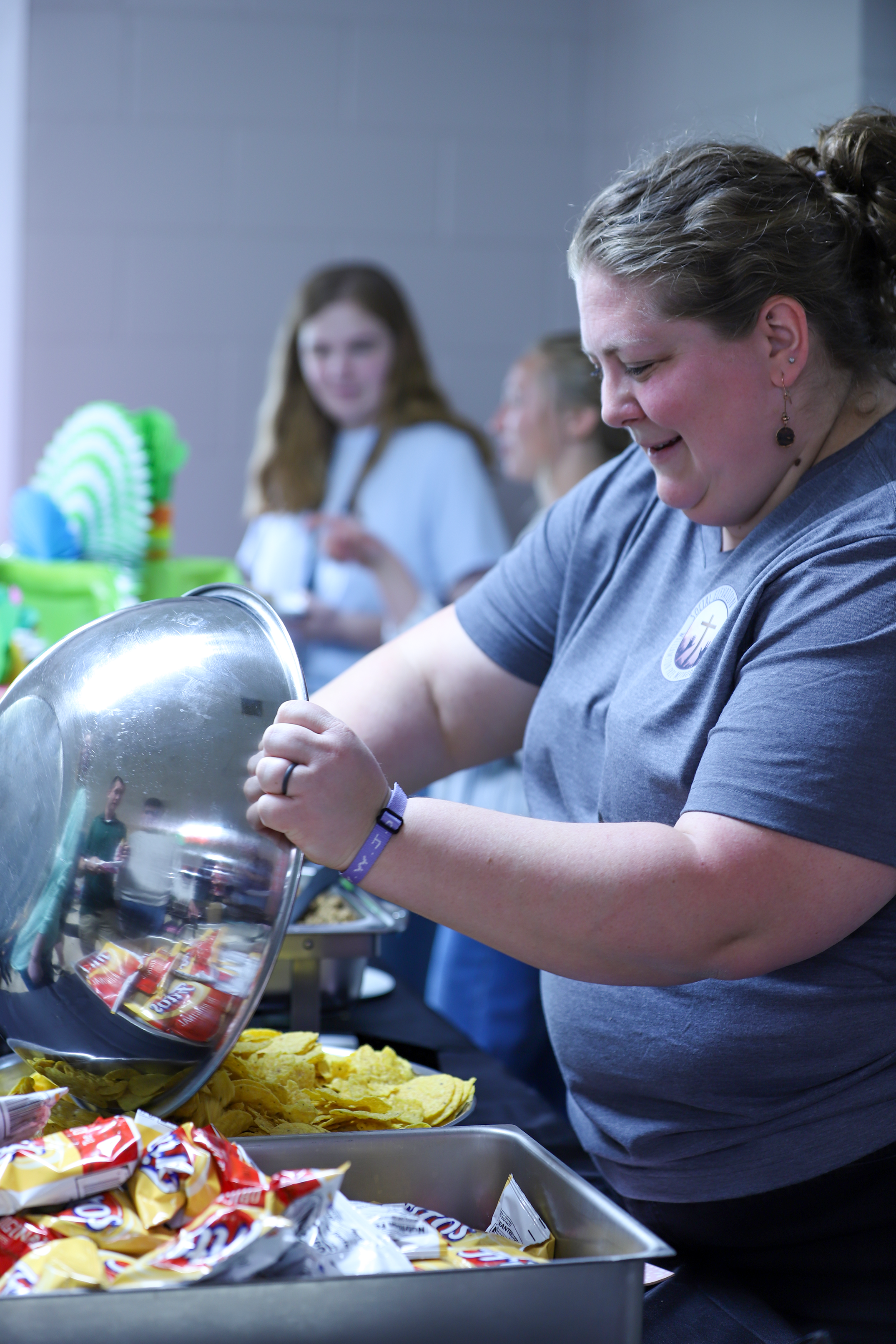 A woman serving snacks at a buffet table during an event, with a woman in a white dress and other guests in the background.
