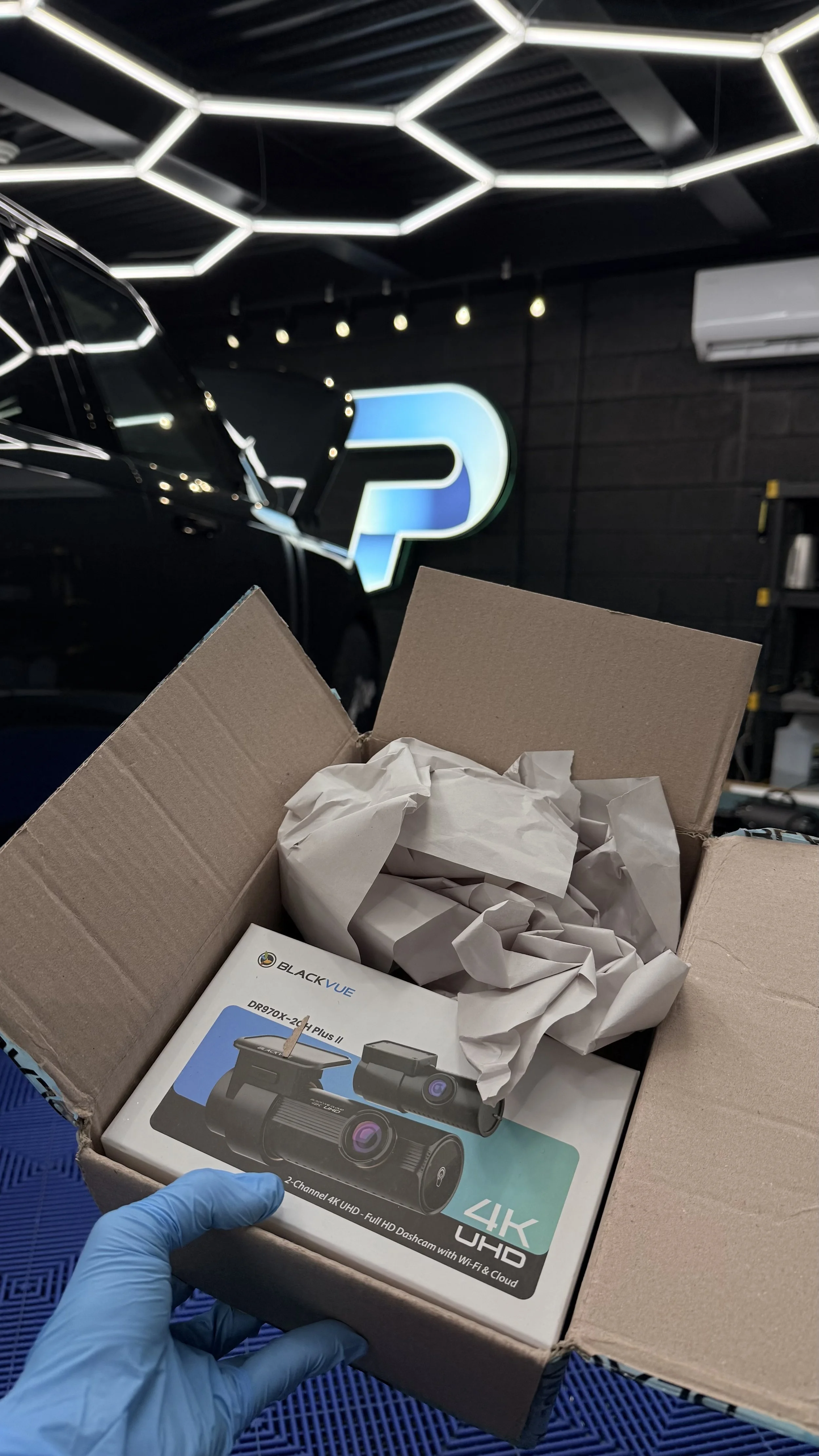 Open cardboard box containing a BLACKVUE DR970X-2CH Plus 4K UHD dash camera and crumpled packing paper, with a person wearing blue gloves holding the box in a modern office or showroom with a black wall, a blue illuminated logo, and contemporary geometric ceiling lights.