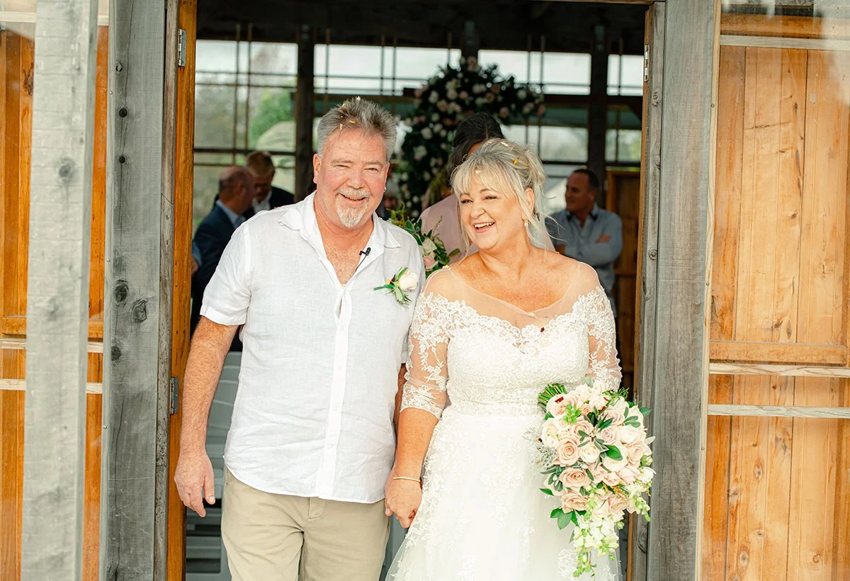 . Andrew Coffey Photographer Weddings
A man in a white shirt and a woman in a wedding dress holding a bouquet of flowers smiling as they walk through a wooden doorway at their wedding.