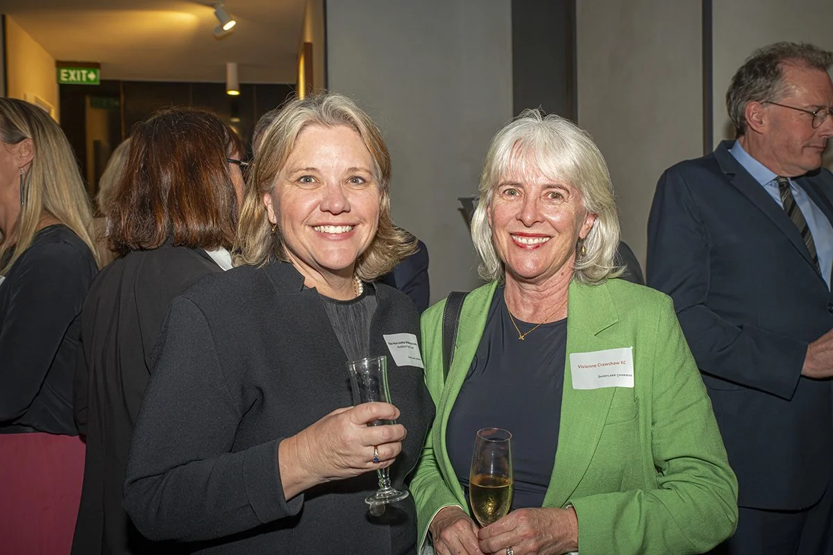 Two women smiling at a social event, holding glasses of wine, standing in a room with other people and an exit sign in the background.
