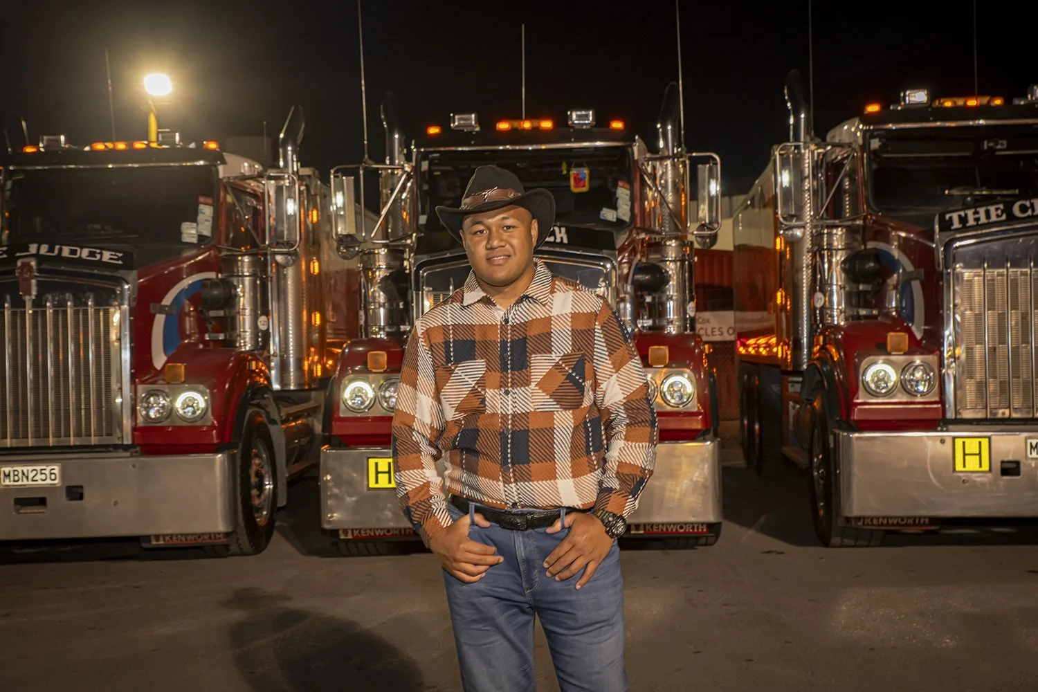 A young man wearing a cowboy hat, checkered shirt, and jeans standing in front of several red fire trucks at night.