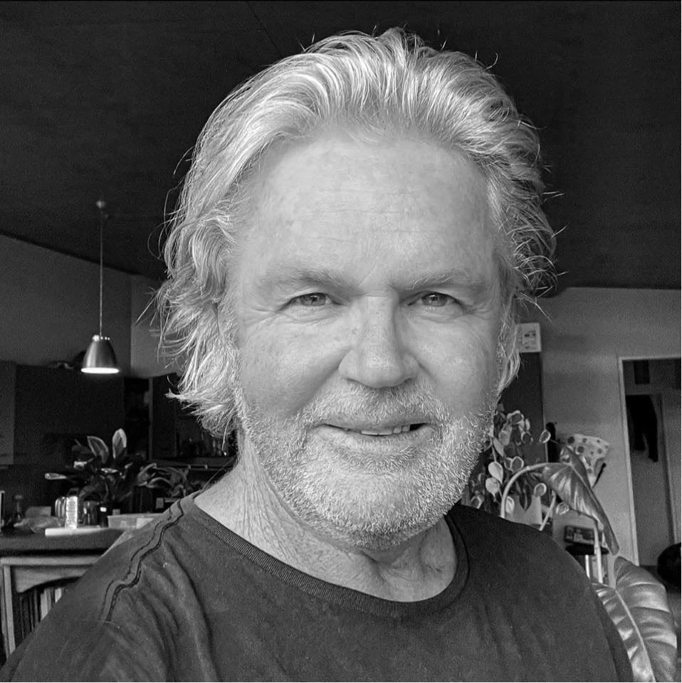 Andrew Coffey photographer About.
Black and white photo of a smiling older man with shoulder-length wavy hair and a beard, indoors with plants and furniture in the background.