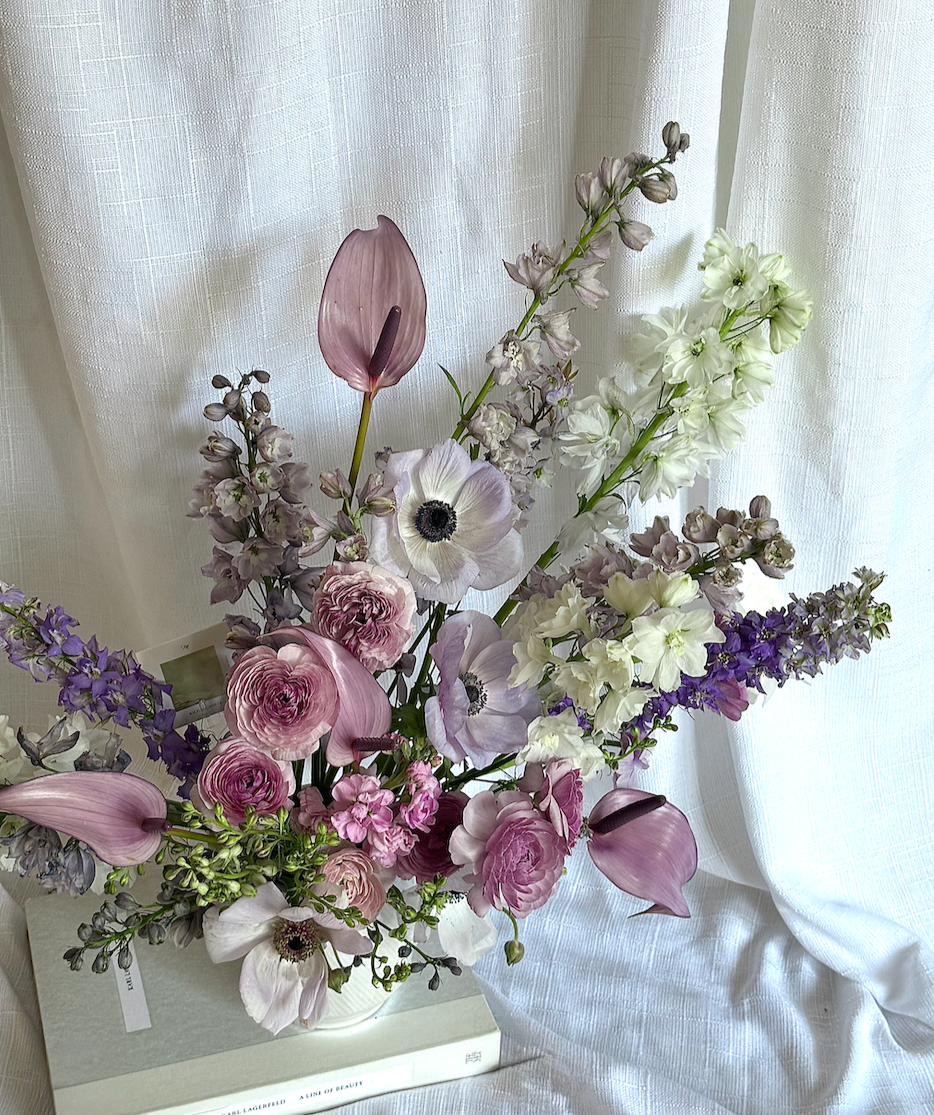 A tall bouquet of pastel-colored flowers in a white vase, including pink calla lilies, anemones, snapdragons, and delphiniums, placed on a white surface with a light curtain backdrop.