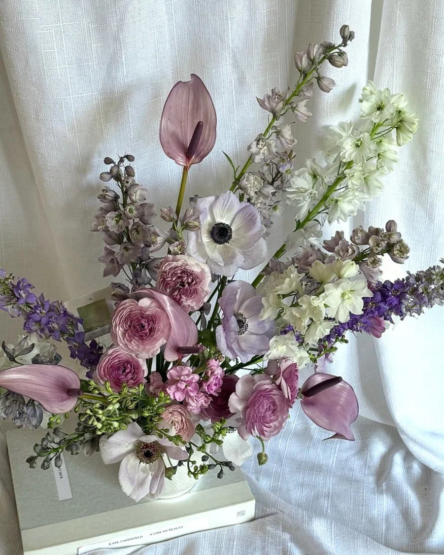 A floral arrangement with pink, purple, and white flowers in a white vase, placed on top of a stack of two books in front of a sheer white curtain.