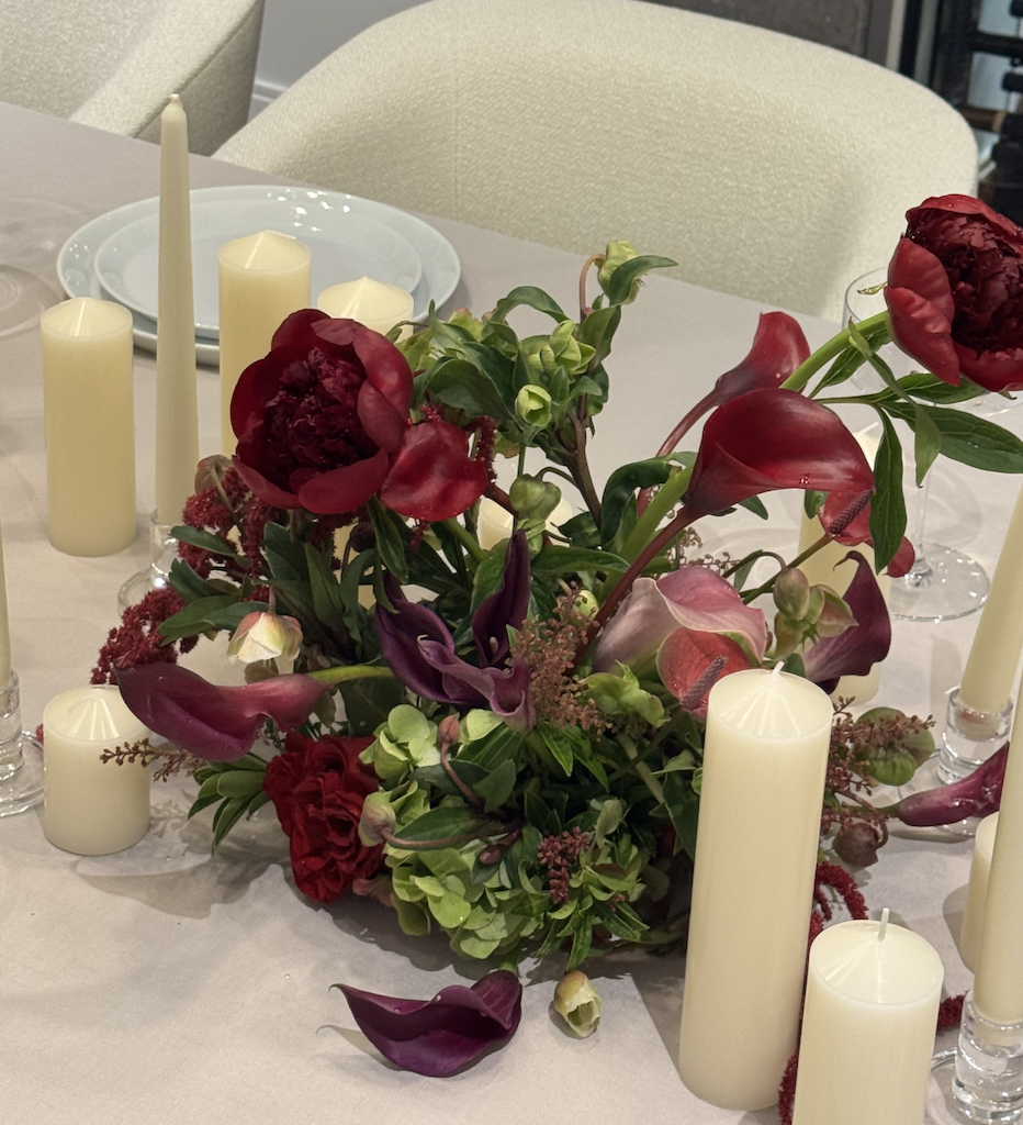 A floral centerpiece with red, pink, and purple flowers surrounded by tall white candles on a dining table.