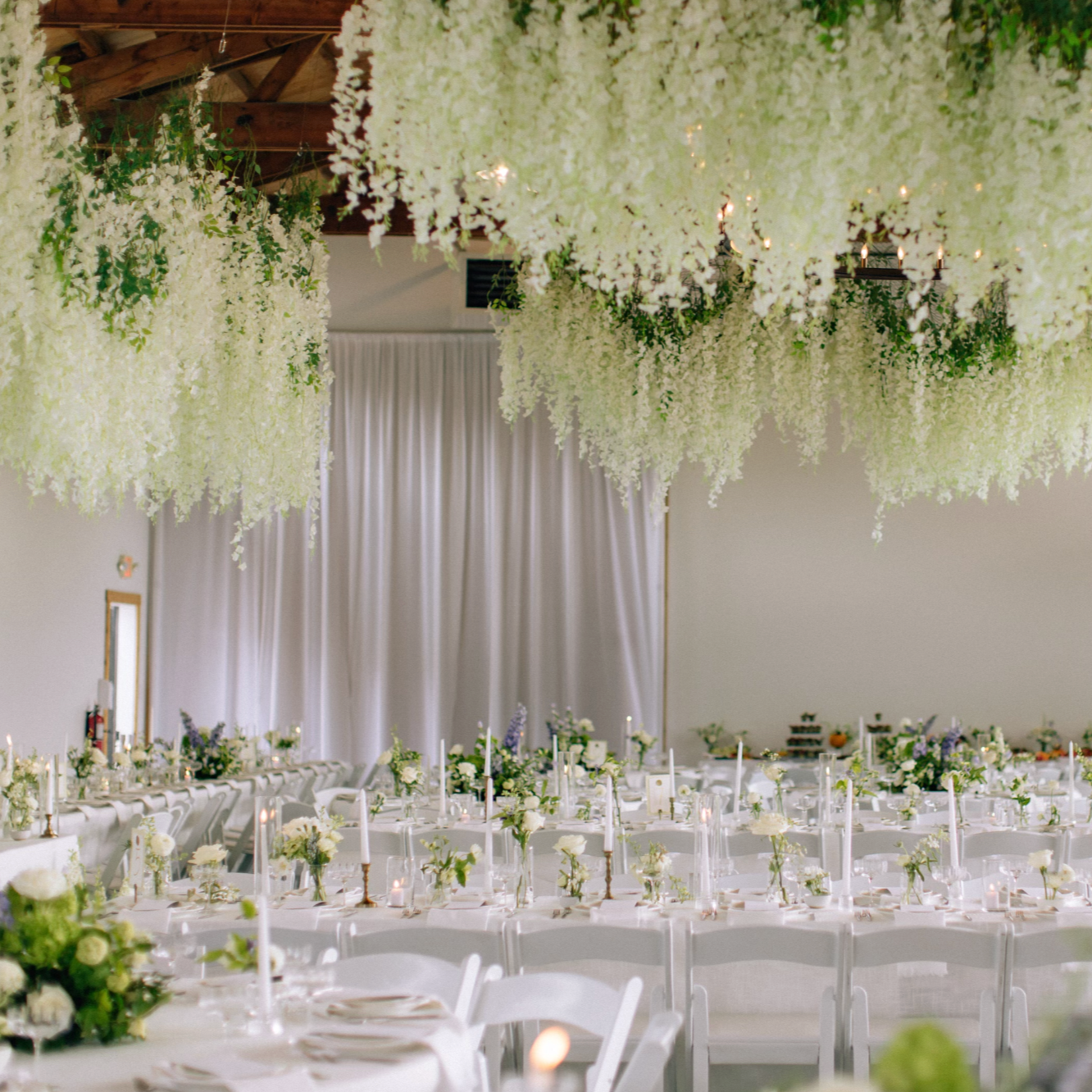 Elegant banquet hall decorated with white flowers and draped curtains, with hanging floral arrangements and many tables adorned with candles and floral centerpieces for a formal event.