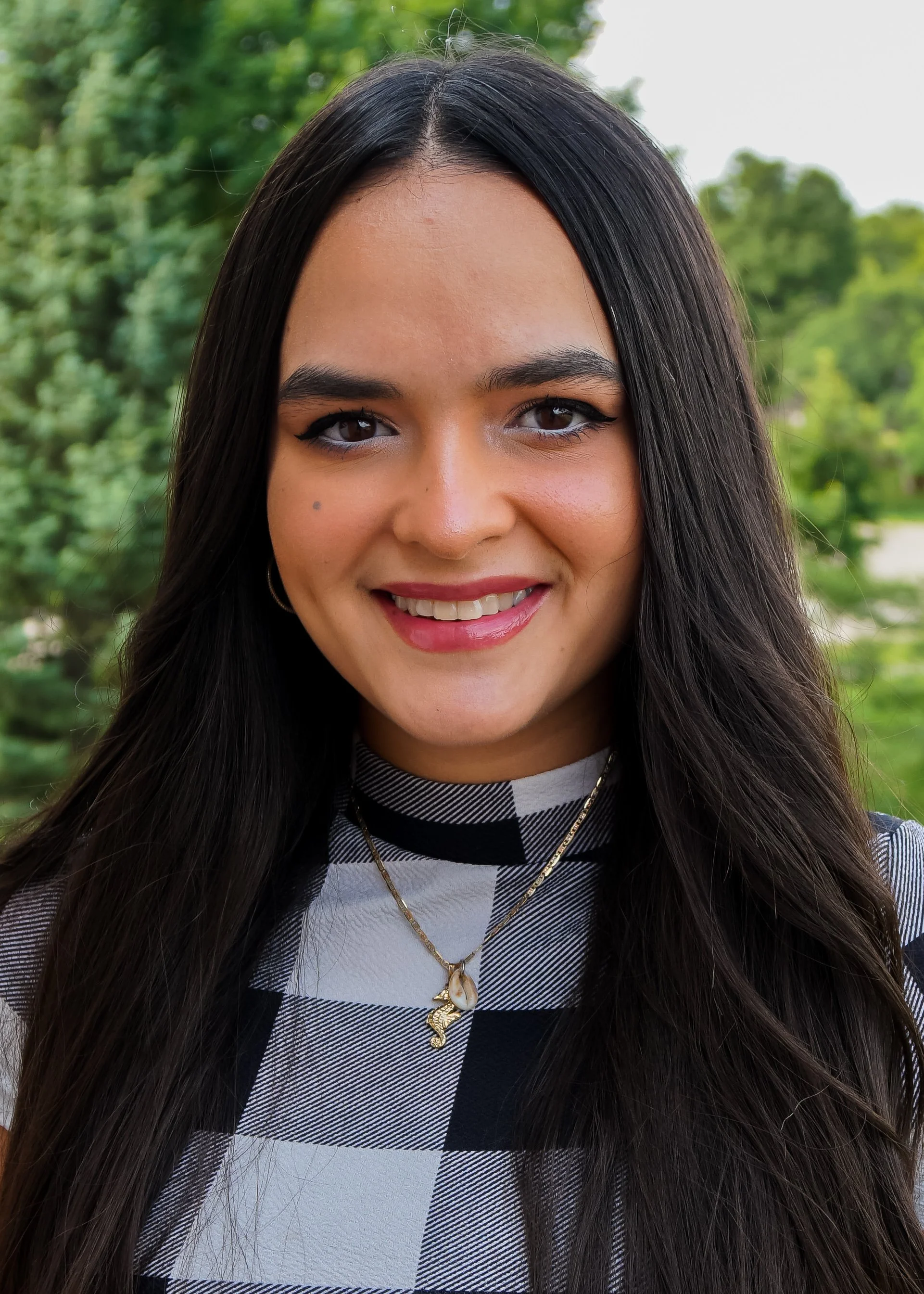 Caroline Cortés Babilonia, Psy.D. A woman with long, dark hair wearing a black-and-white patterned shirt and gold necklace. Behind her is a green wooded background.