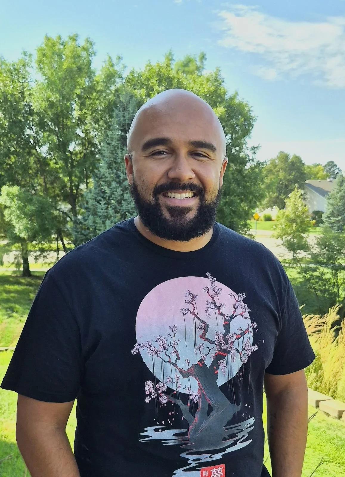 Kashawn Lungelow. A man with a beard and wearing a black shirt. There is a green background behind him with trees and grass.