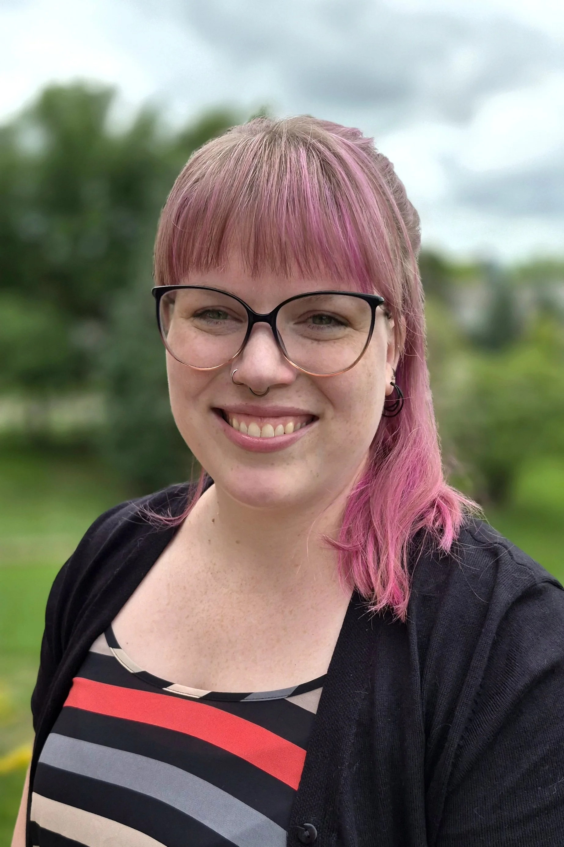 Picture of Alexandra Bissell, she has light hair with bright pink highlights, glasses, and she is wearing a black sweater and striped shirt.
