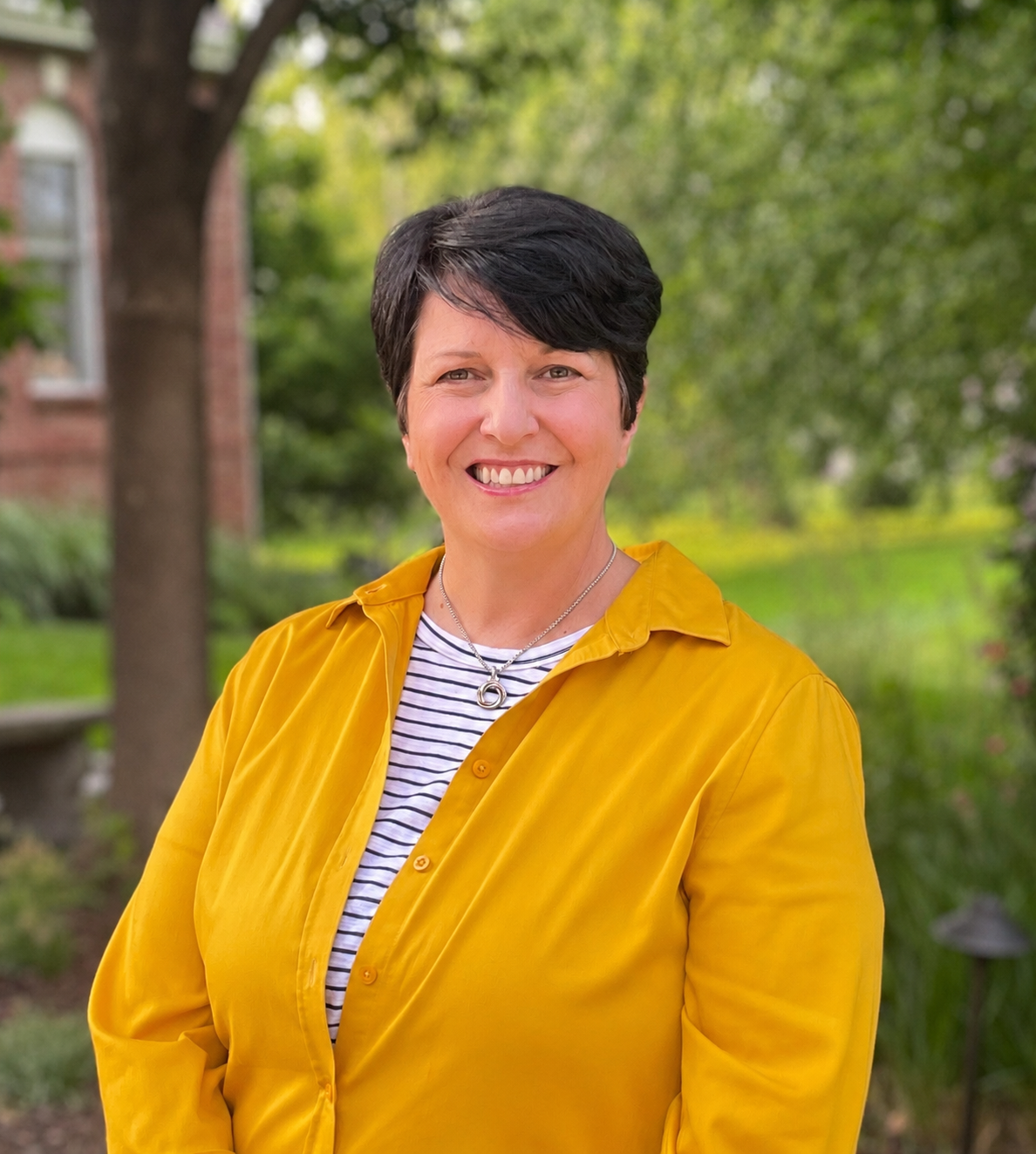 A woman with short-cropped dark hair, smiling. She is wearing a yellow shirt with a white shirt underneath, the shirt has horizontal black stripes. She is standing in front of a woodsy setting with trees and shrubs and a red brick house.