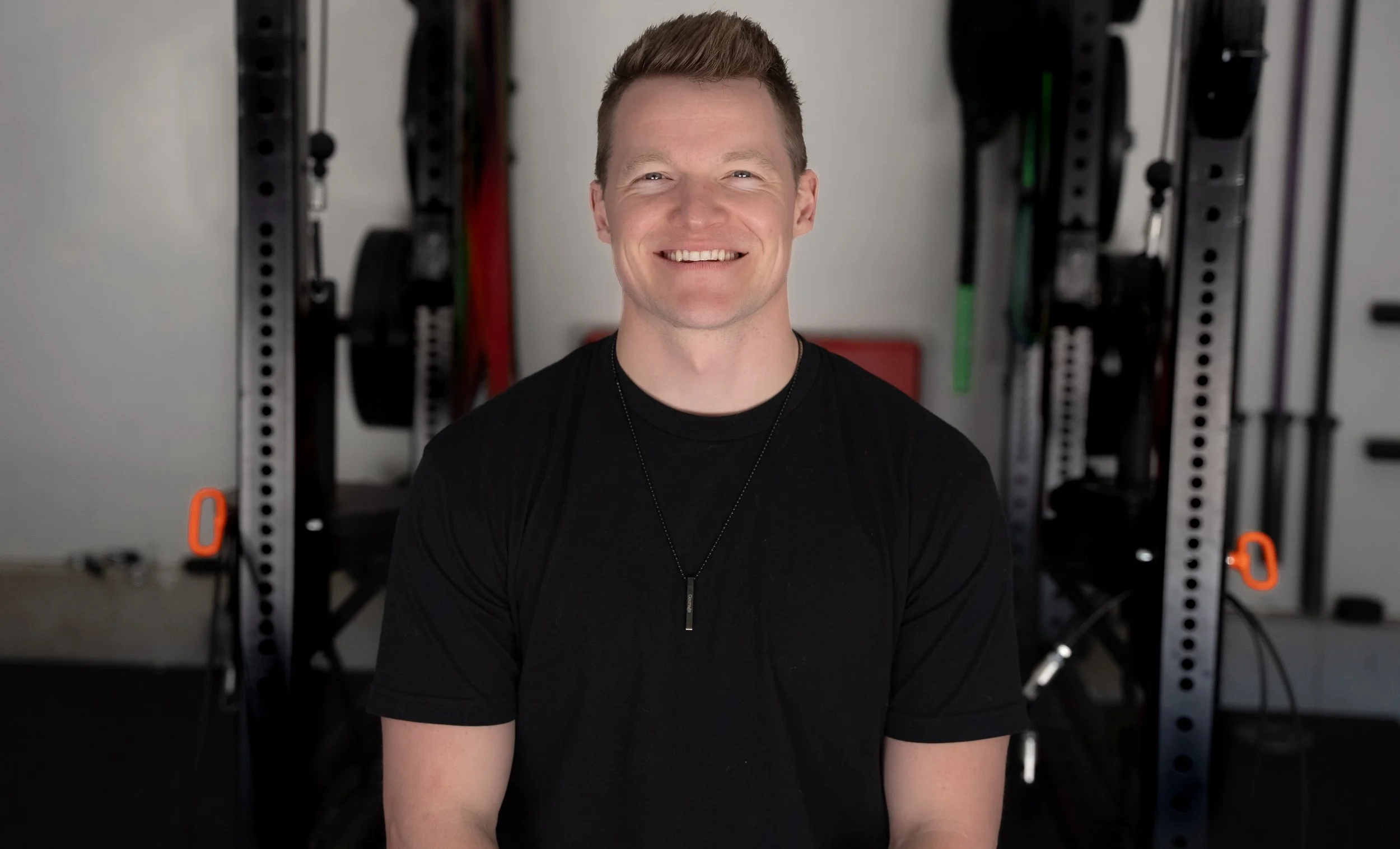 A young man with short brown hair, wearing a black t-shirt and a necklace, smiling at the camera in a gym or workout space with exercise equipment in the background.