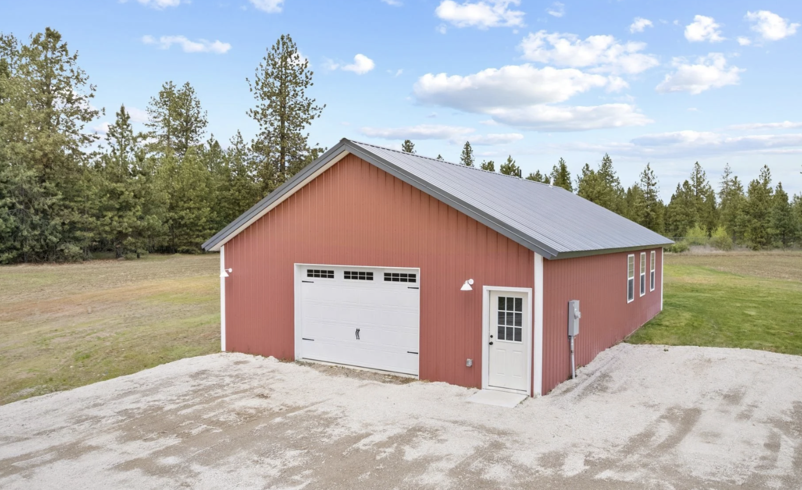 Red barn with a gray metal roof, white garage door, and side door, set on a gravel driveway with a grassy area, surrounded by trees and a partly cloudy sky.