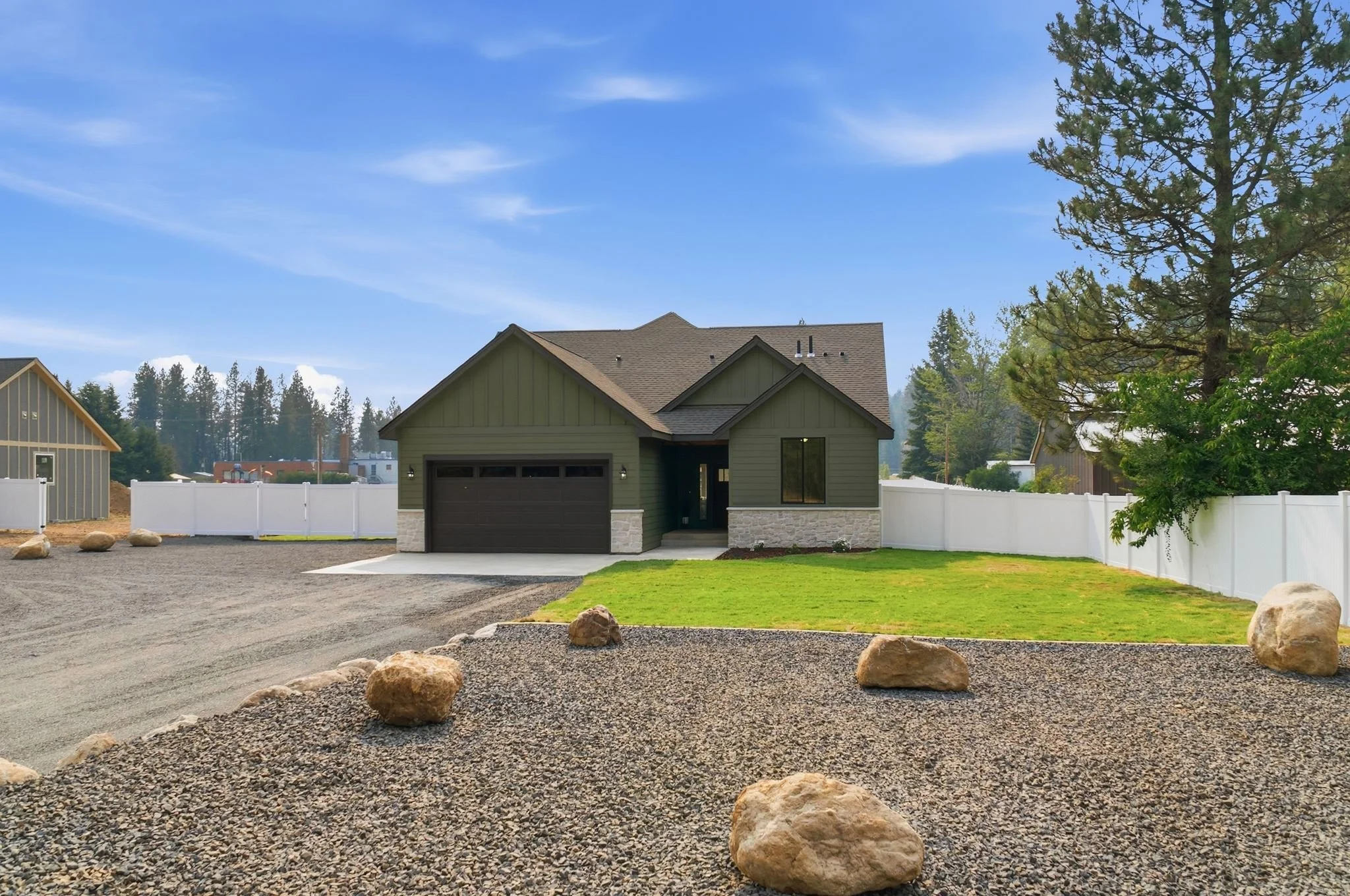 A newly built house with green exterior walls, a dark garage door, and a small front porch, surrounded by a white fence and a gravel yard with large rocks, trees, and a blue sky overhead.