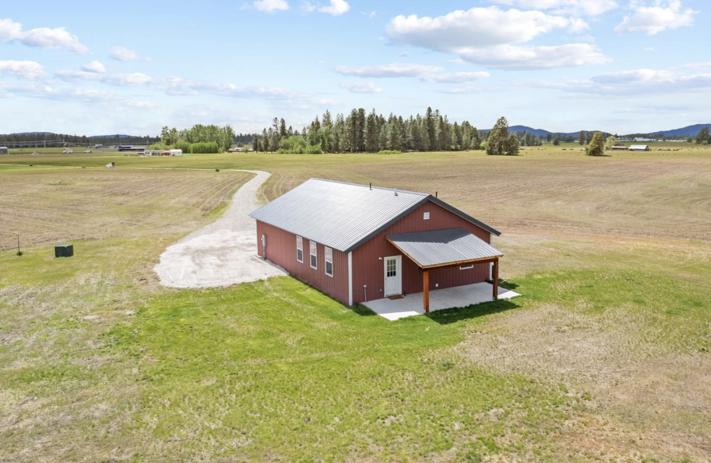 A red barn with a metal roof located in a rural field with green grass, surrounded by open farmland and distant trees under partly cloudy sky.