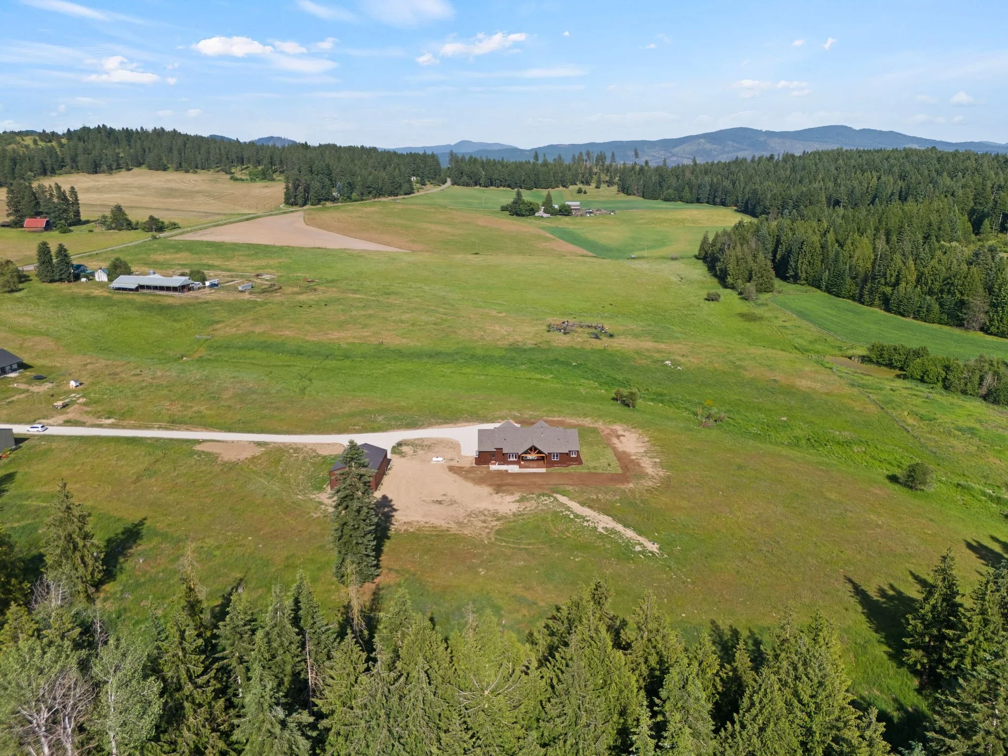 Aerial view of a rural landscape with green fields, scattered farmhouses, large forests, and mountain ridges in the distance under a partly cloudy sky.