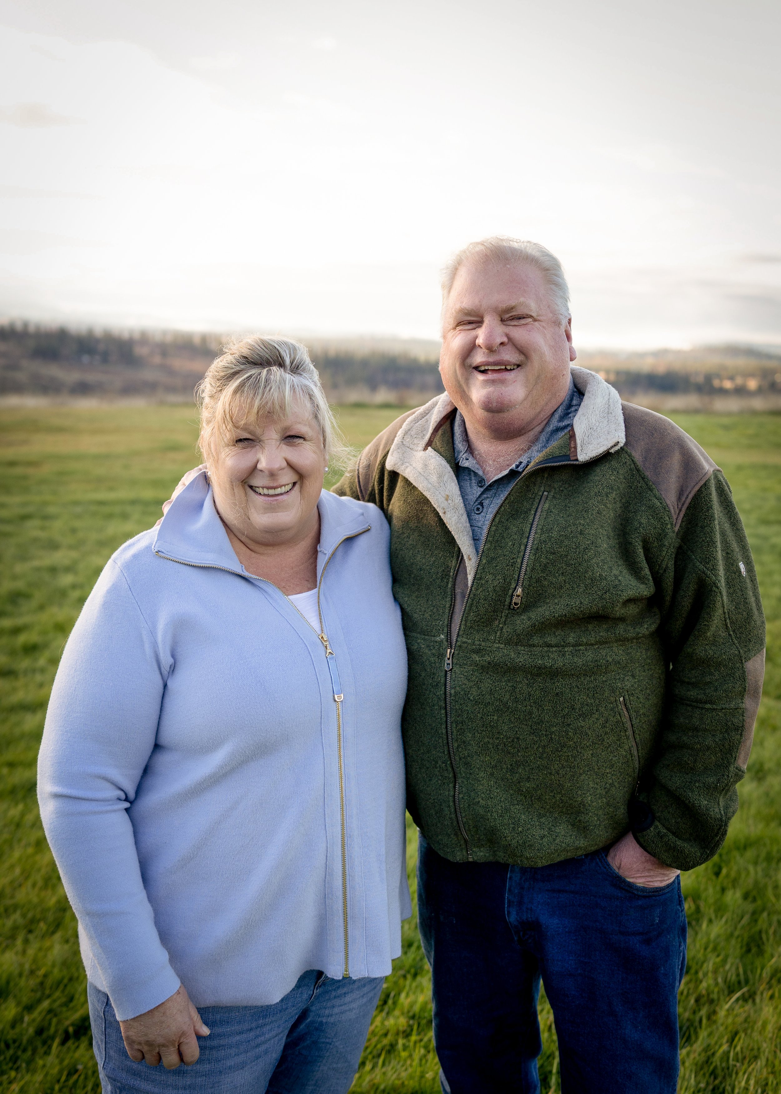 A smiling elderly woman and a smiling elderly man standing outdoors in a grassy field during daytime. The woman is wearing a light blue zip-up jacket and the man is wearing a green and brown fleece jacket. They are close together, with their shoulders touching, against a background of open fields and a partly cloudy sky.