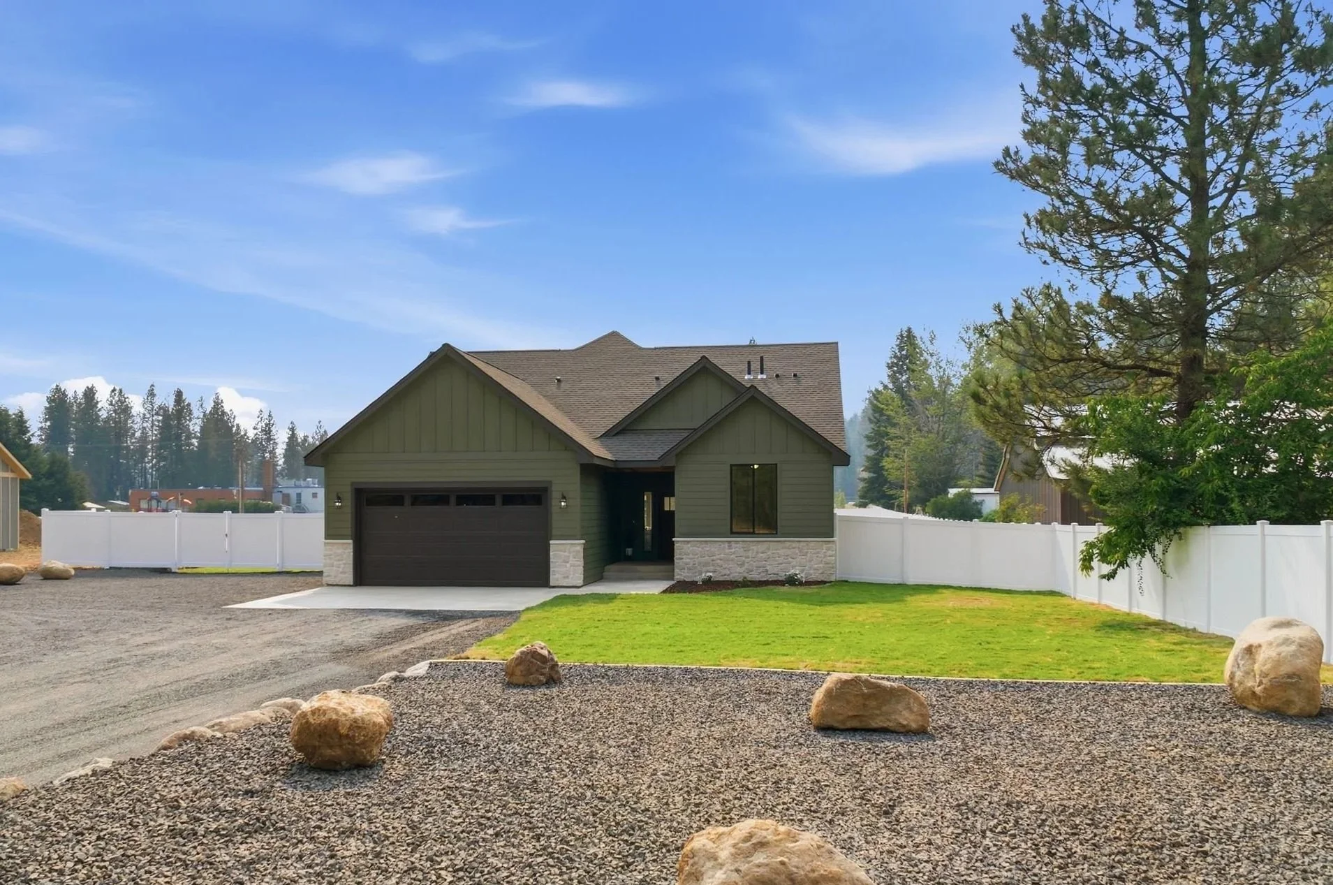 Newly built house with a gray garage door, stone accents, and dark green siding, surrounded by a white fence, with a gravel driveway and rocks in the foreground, tall trees, and a bright blue sky.