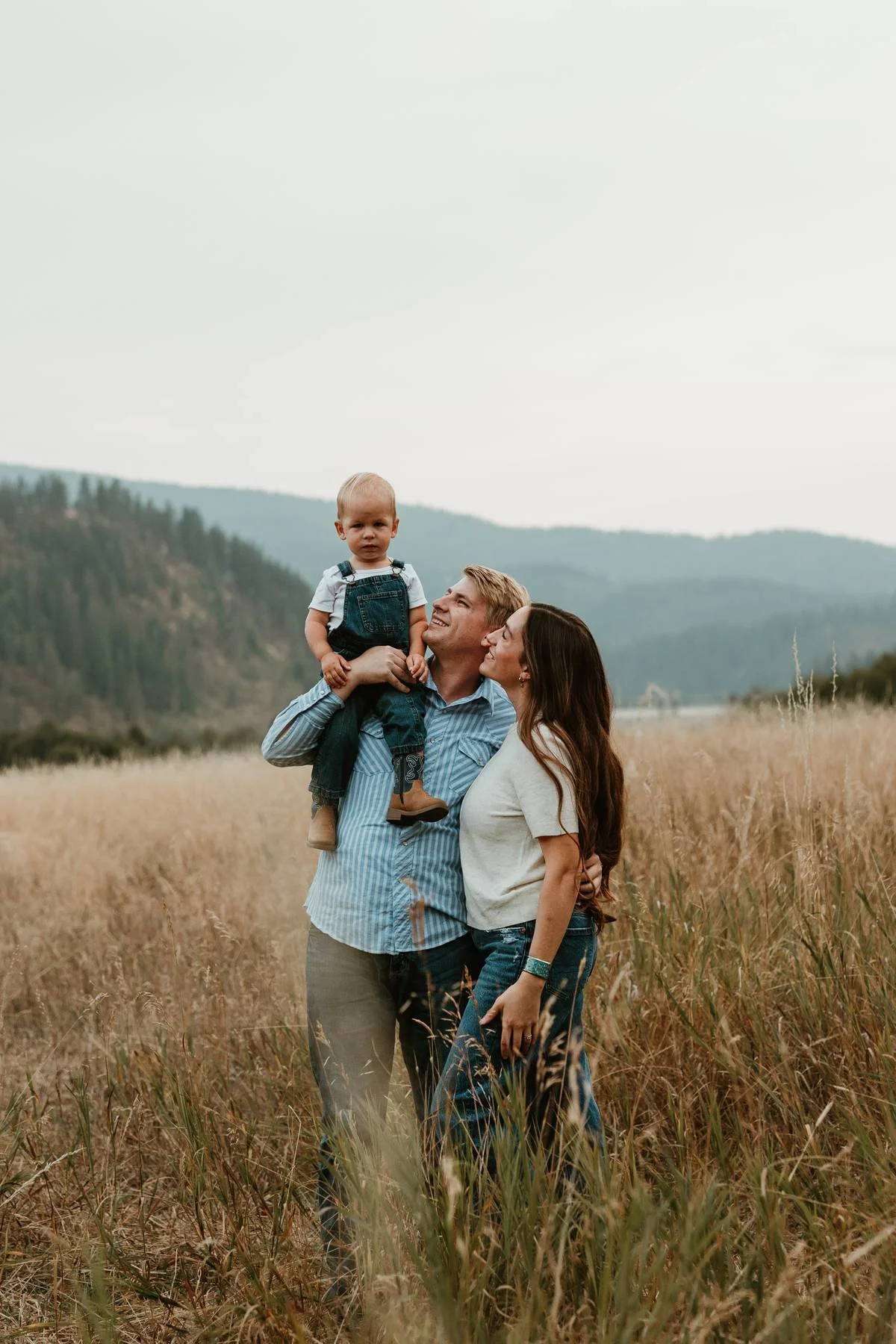 A family of three standing in a field with tall grass, with mountains and cloudy sky in the background. The father is holding their young child on his shoulder, and the mother is standing close, all looking happy.
