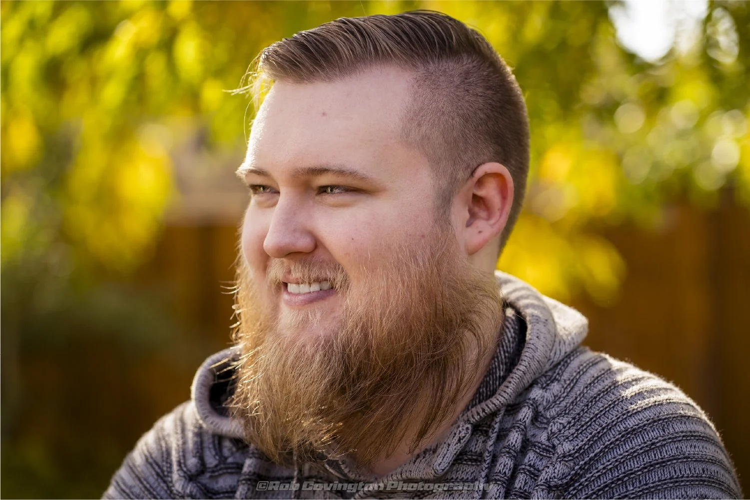 Portrait photography by Rob Covington. A smiling man with a full beard and short hair, wearing a gray patterned hoodie, outdoors with yellow-green blurred foliage in the background.