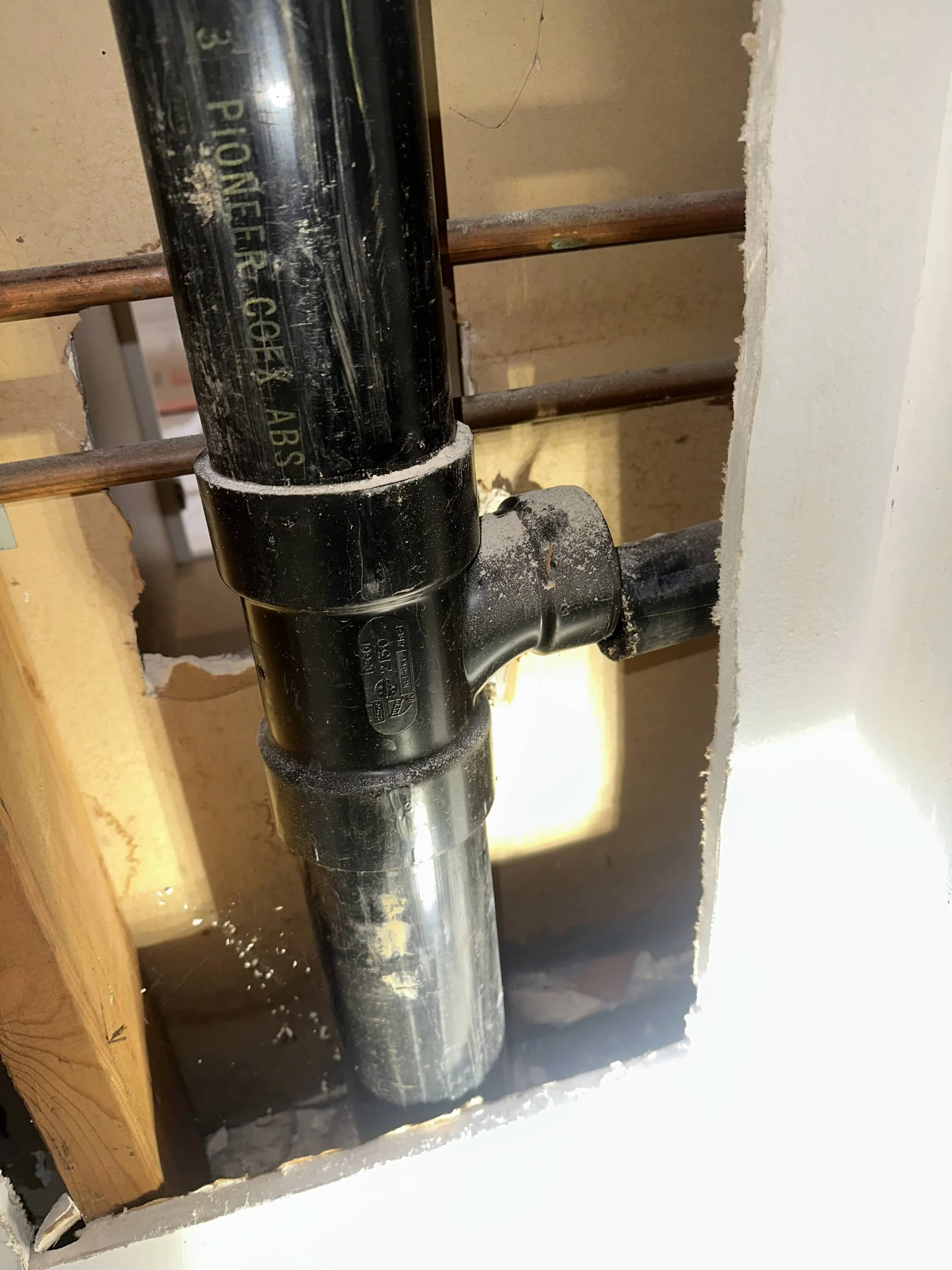 A close-up of black plumbing pipes, including a T-joint, inside a partially open wall. The surrounding area shows wooden studs and exposed drywall. Sunlight and dust are visible.