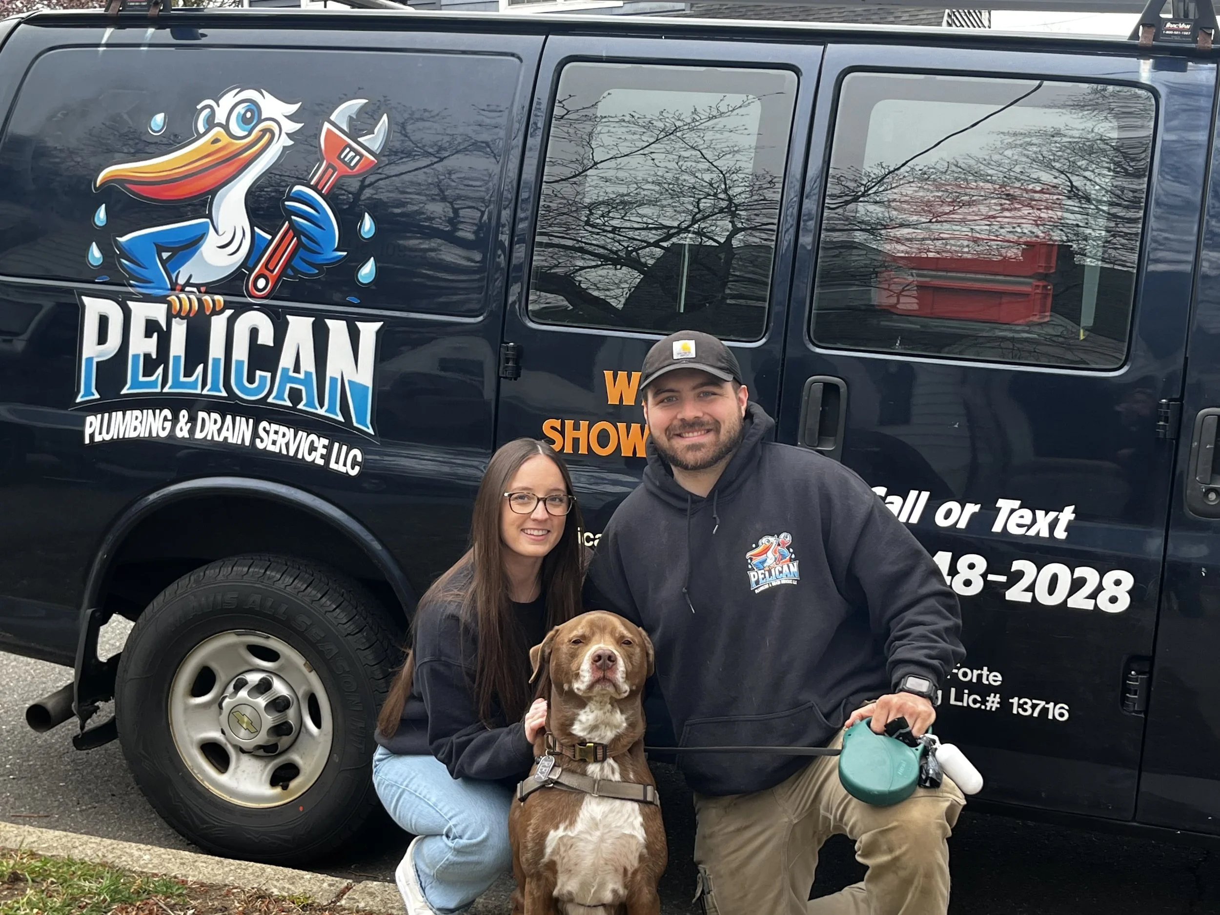 Two people kneeling next to a brown dog in front of a dark blue van with "Pelican Plumbing & Drain Service LLC" logo, featuring a cartoon pelican holding a wrench. The van also displays the company's text and contact information.