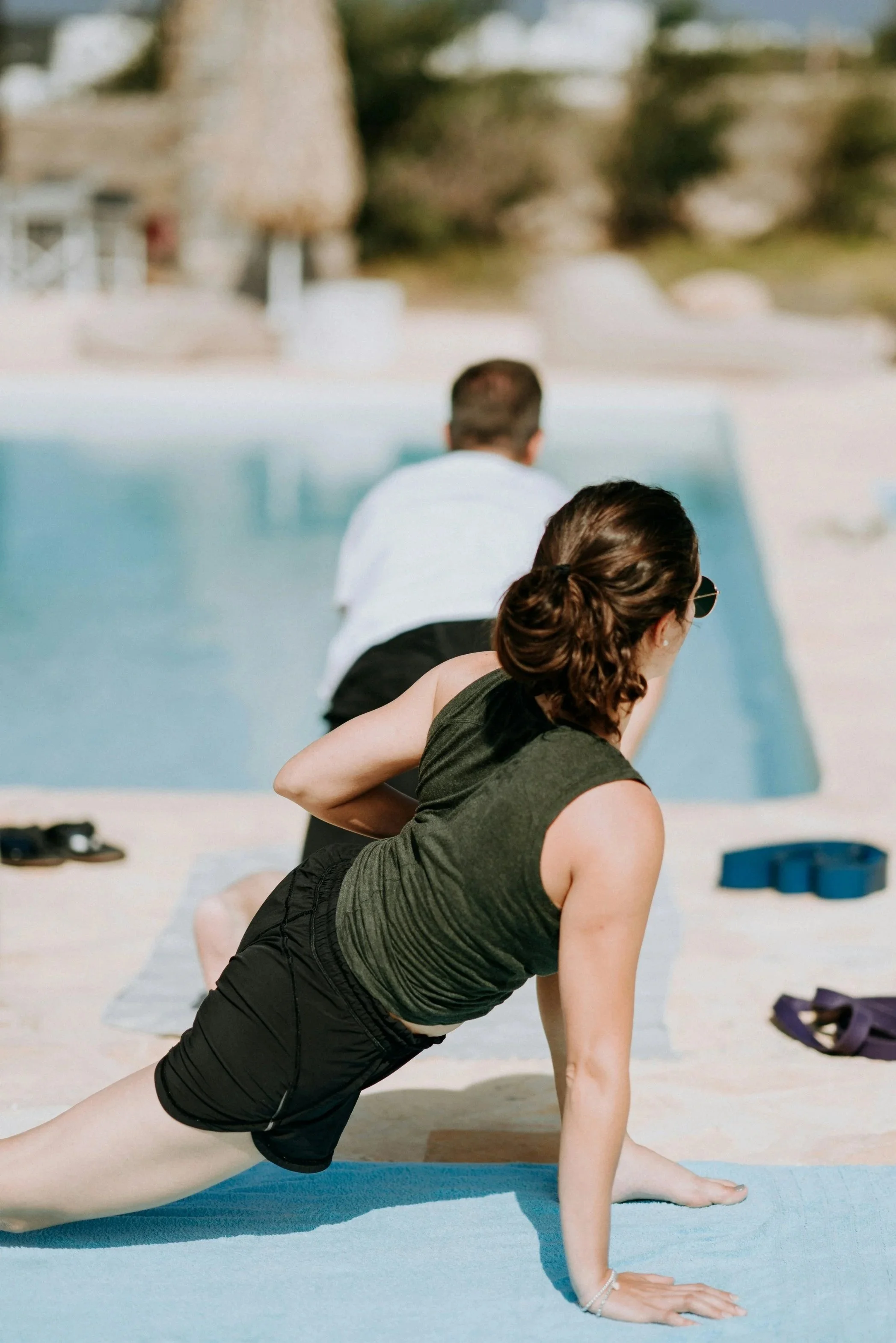 A woman practicing yoga outdoors near a pool, with a man sitting in the background.
