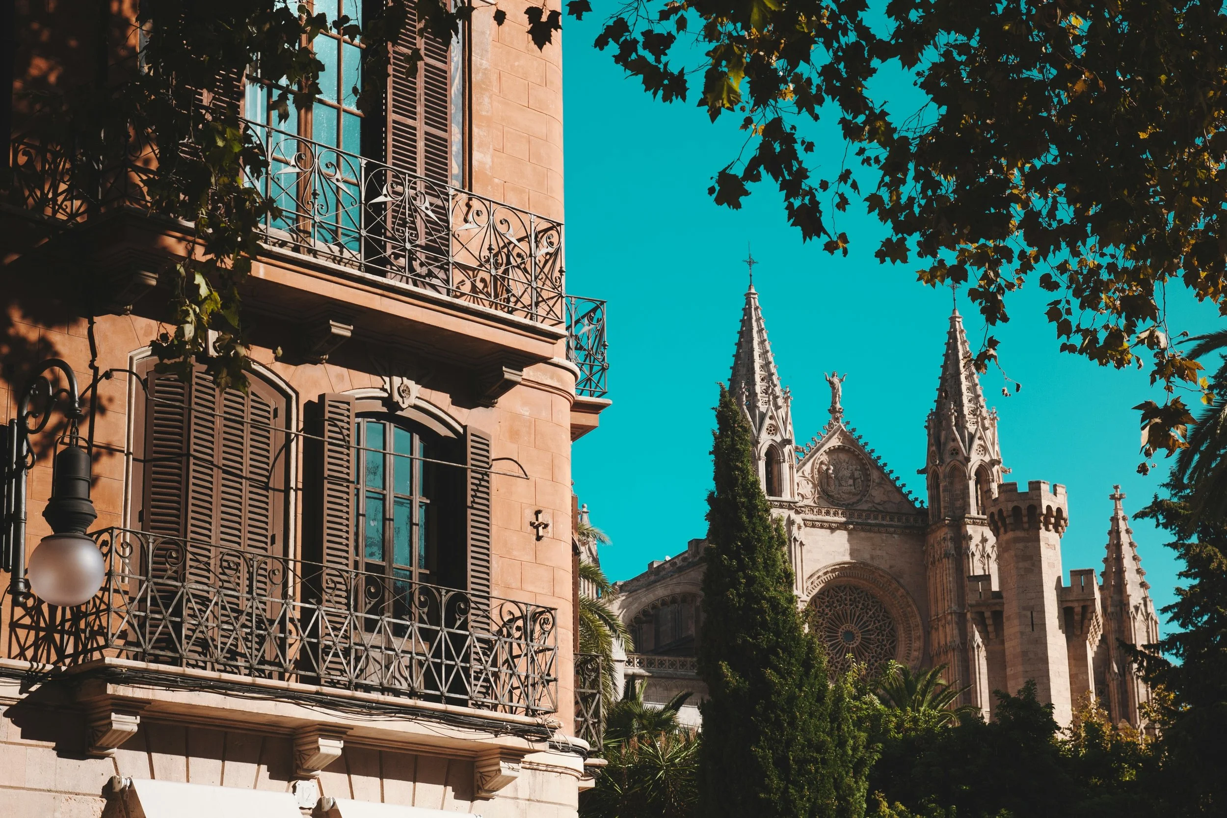 A European city street scene showing a residential building with wrought iron balconies and shutters, and a Gothic cathedral with spires and rose window, surrounded by trees under a bright blue sky.