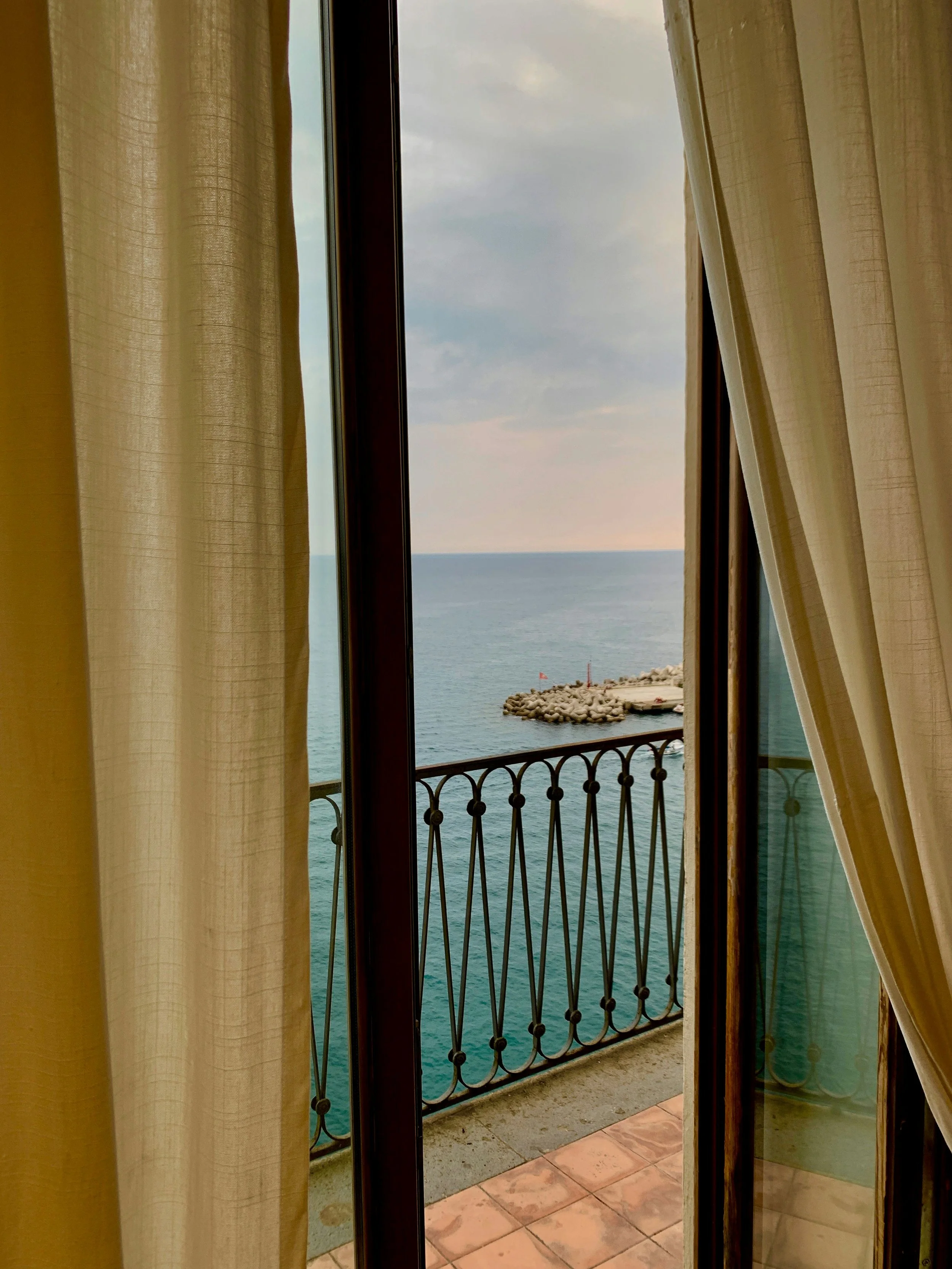View of the ocean through an open sliding glass door with cream curtains, showing a small pier or breakwater with a flag and a red lighthouse in the distance.