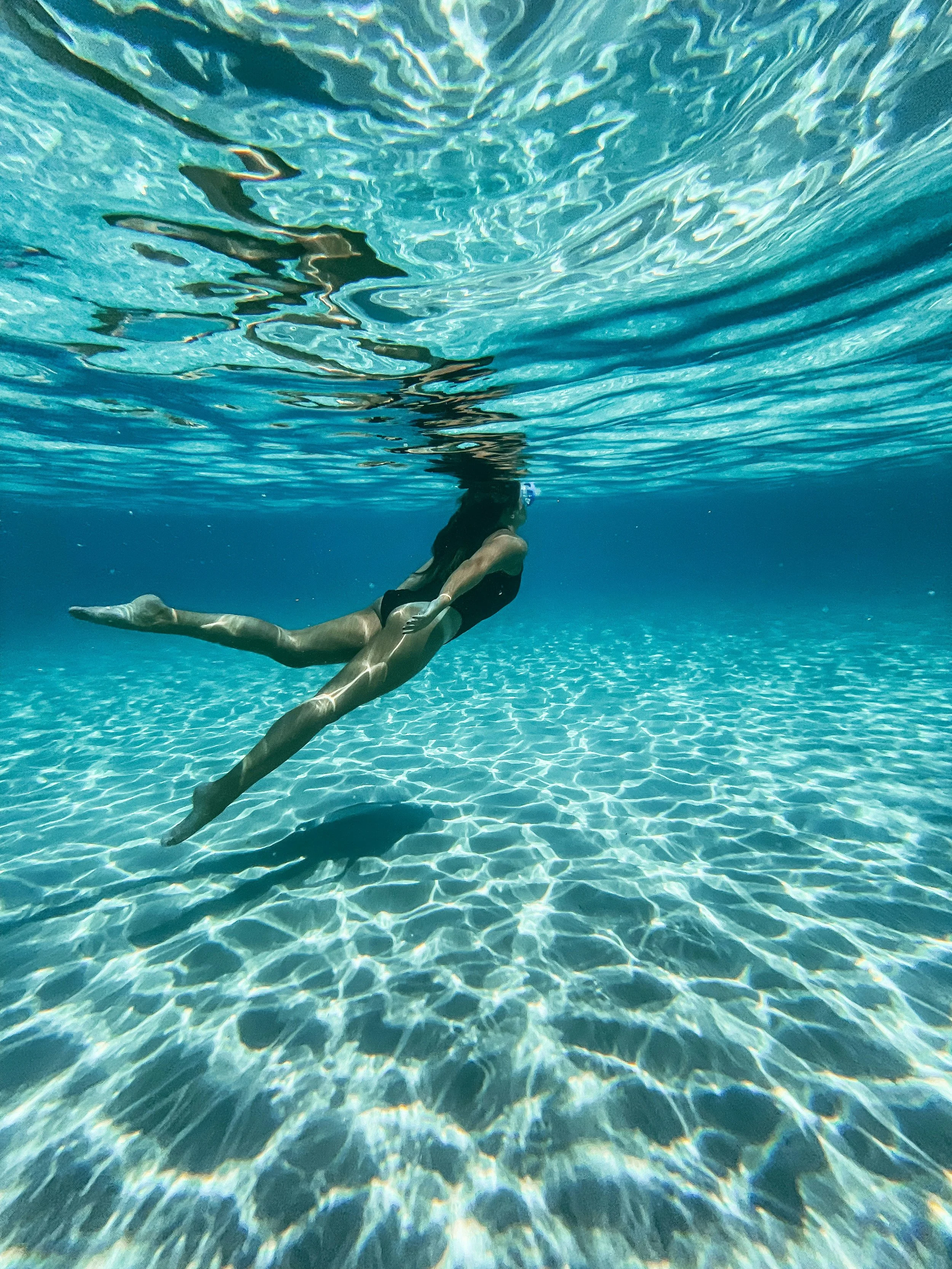 An underwater scene showing a person swimming in a pool, viewed from below. The swimmer is in black swimwear, and sunlight creates rippling reflections on the pool floor.