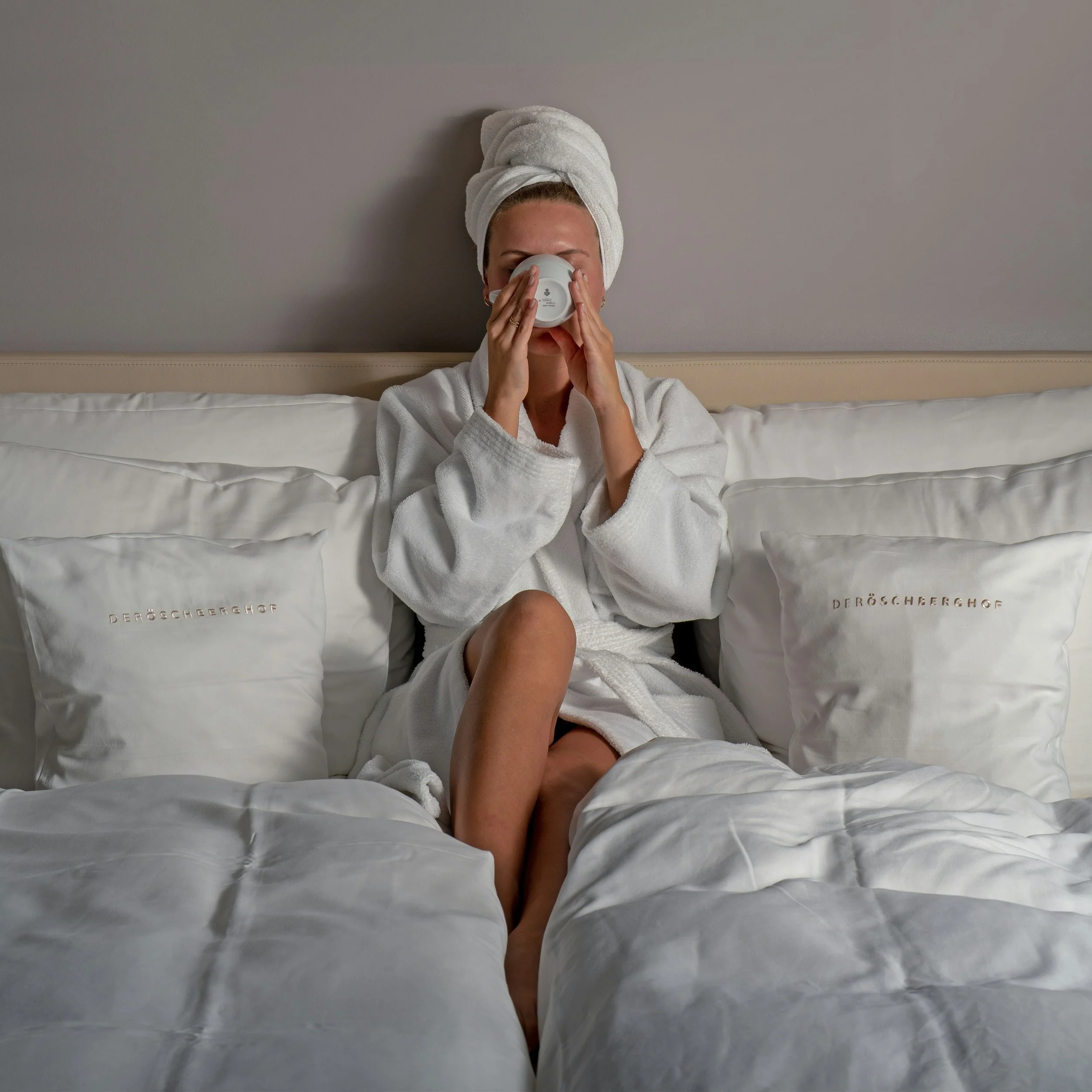 A woman in a white bathrobe and towel wrapped around her head sits on a bed, drinking from a white mug with her eyes closed.