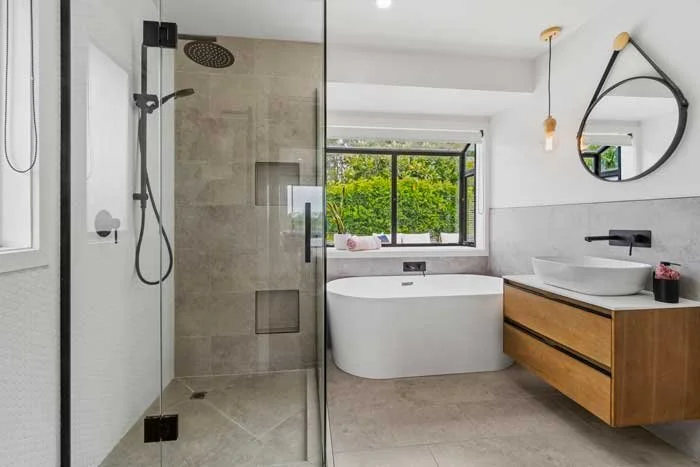 Modern bathroom featuring a glass shower with black fixtures, a freestanding bathtub near large windows, a wooden vanity with a white vessel sink, and a round mirror.