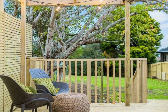 Outdoor deck with wicker chairs and a round ottoman, surrounded by wooden slats, overlooking a yard with a large tree and fence.